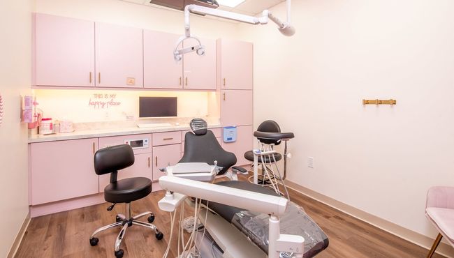 An exam room with a dental chair, medical equipment, and black stools against light pink cabinetry and wood flooring.