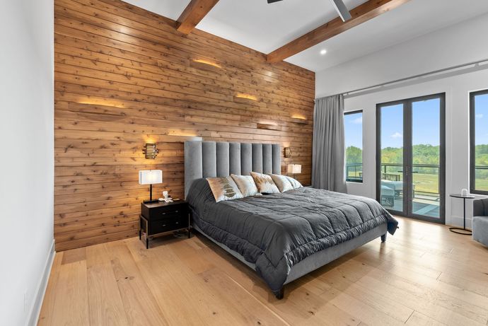 Bedroom with wood accent wall, gray bed, and large windows overlooking a landscape.