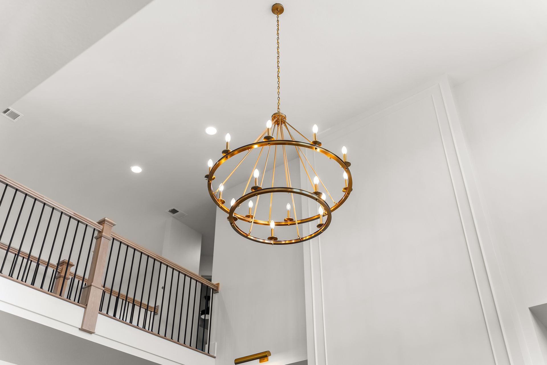 Gold chandelier hanging from a high white ceiling in a room with a second-story balcony.