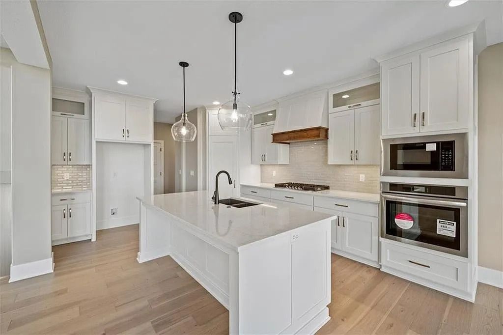 White kitchen with island, cabinets, and built-in appliances. Wooden floor and light fixtures.