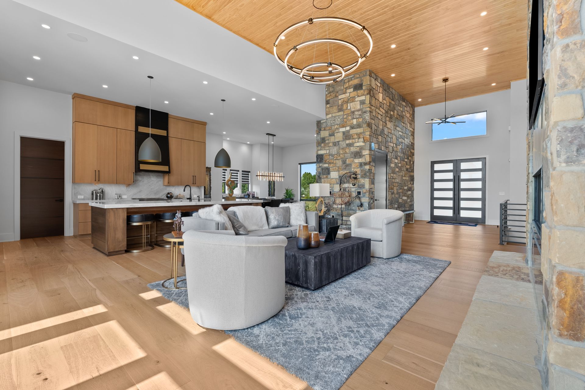 Modern living room with wood floors, stone fireplace, kitchen, and light-filled open space.