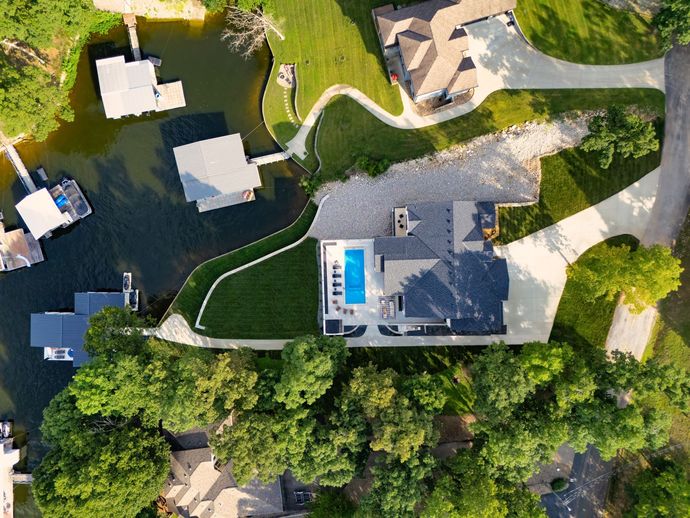 Aerial view of a lakeside house with a pool, docks, and surrounding green trees and grass.