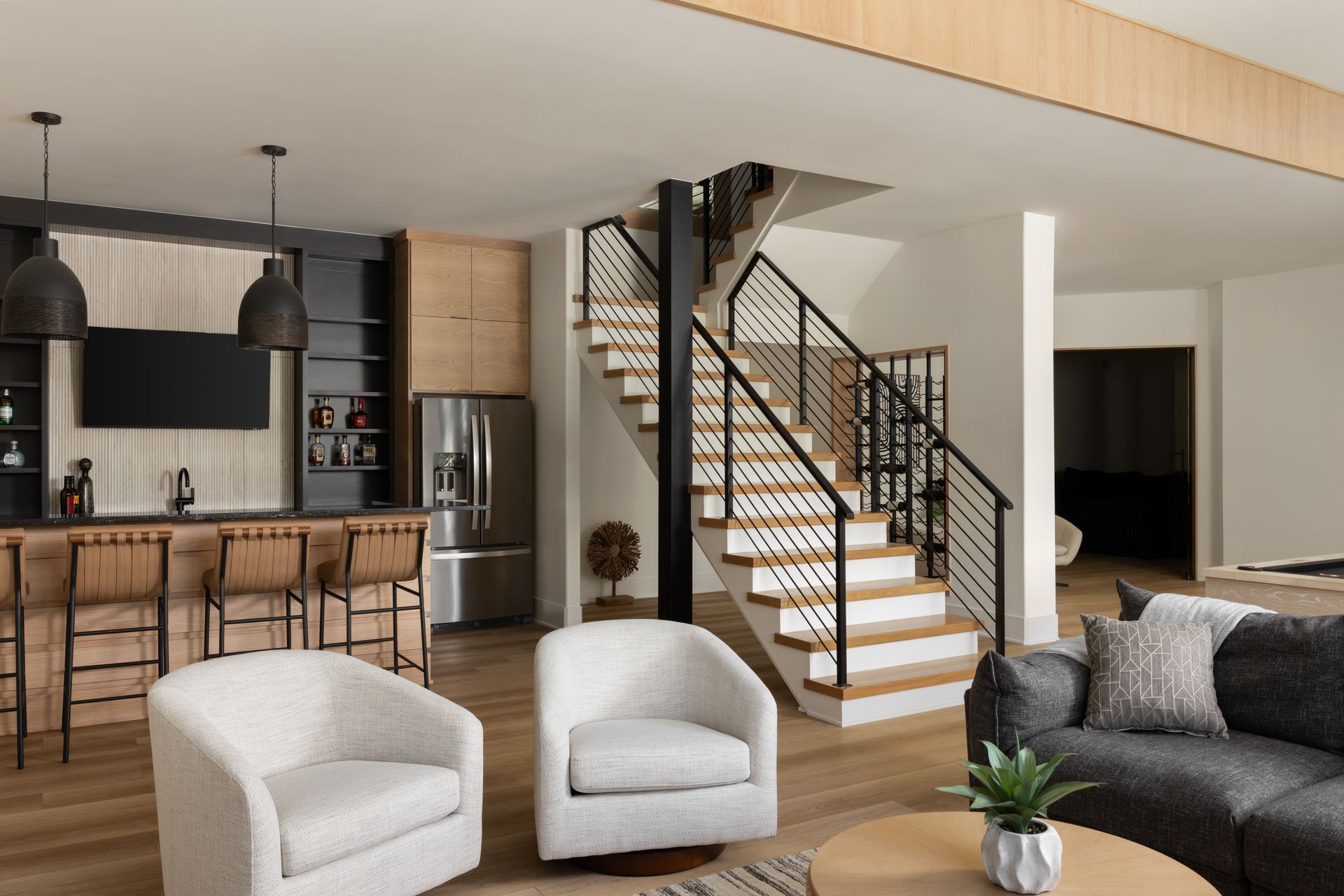 Modern basement with bar, staircase, and seating area; neutral tones, natural wood, and black accents.