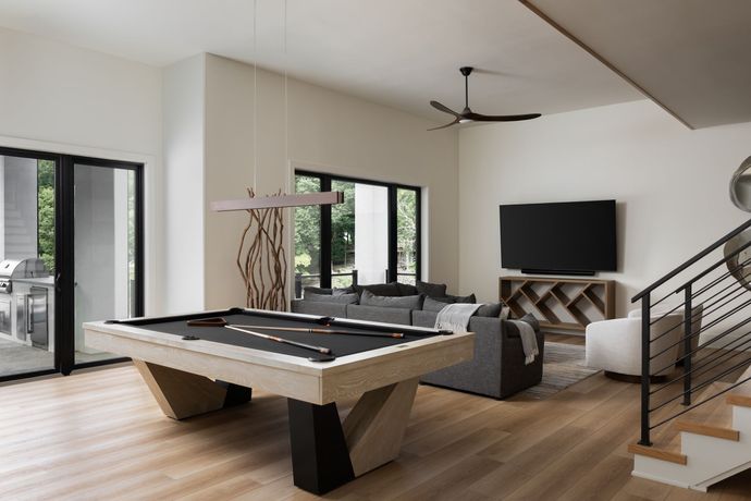 Modern game room with pool table, sofa, TV, and staircase. Light wood floors, neutral walls.