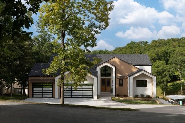 Modern two-story house with a glass garage door, neutral colors, and surrounding trees under a blue sky.