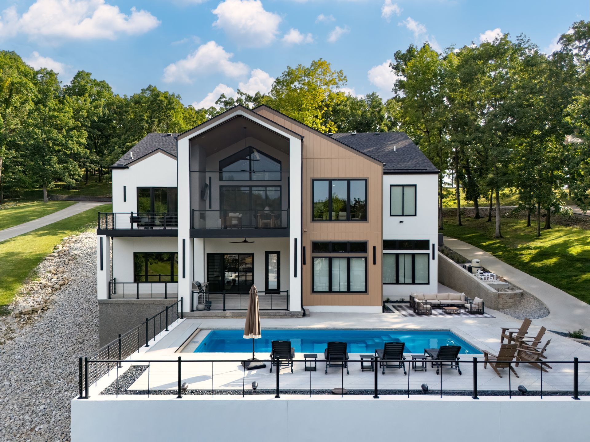 Modern house with pool. White and tan exterior. Black framed windows, deck, and railings. Overlooks lush green trees.