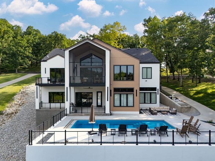 Modern house with pool. White and tan exterior. Black framed windows, deck, and railings. Overlooks lush green trees.