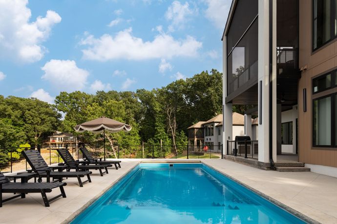 A rectangular pool with lounge chairs, beside a modern house, set against a tree-filled landscape.