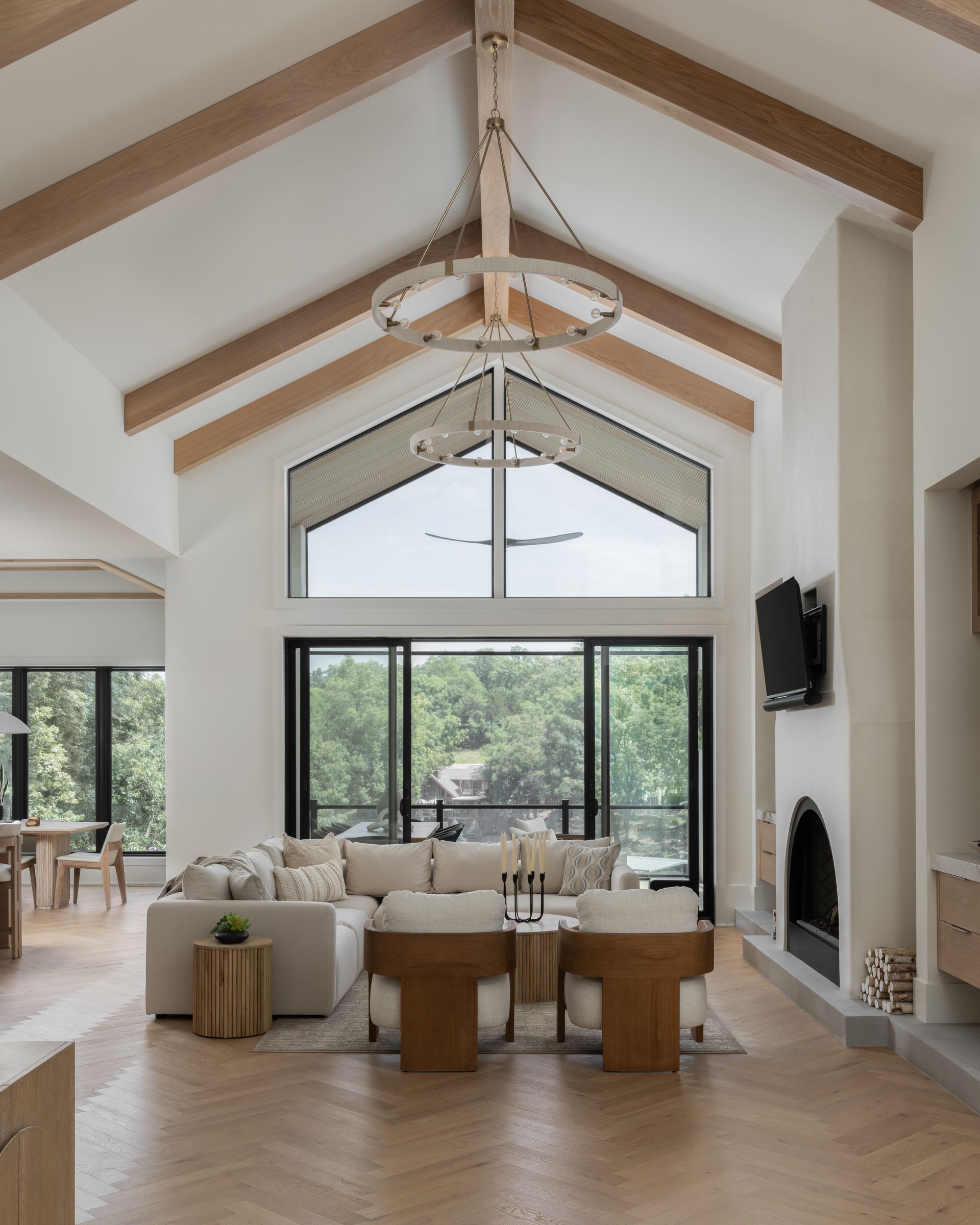 Living room with vaulted ceiling, exposed beams, and large windows overlooking greenery. Beige and white interior with fireplace.