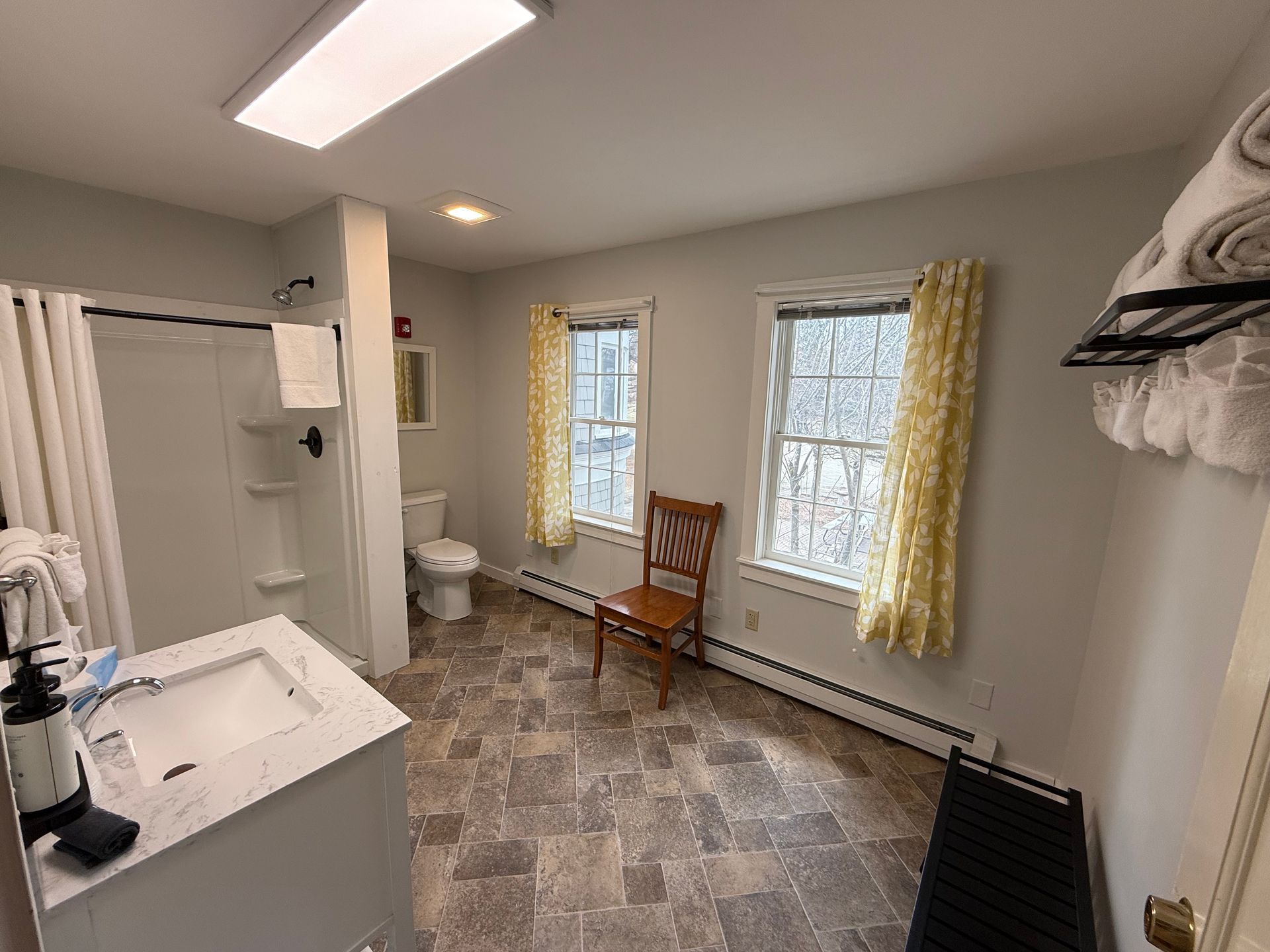 Small bathroom with vanity, toilet, two windows, wooden chair, and tiled floor.