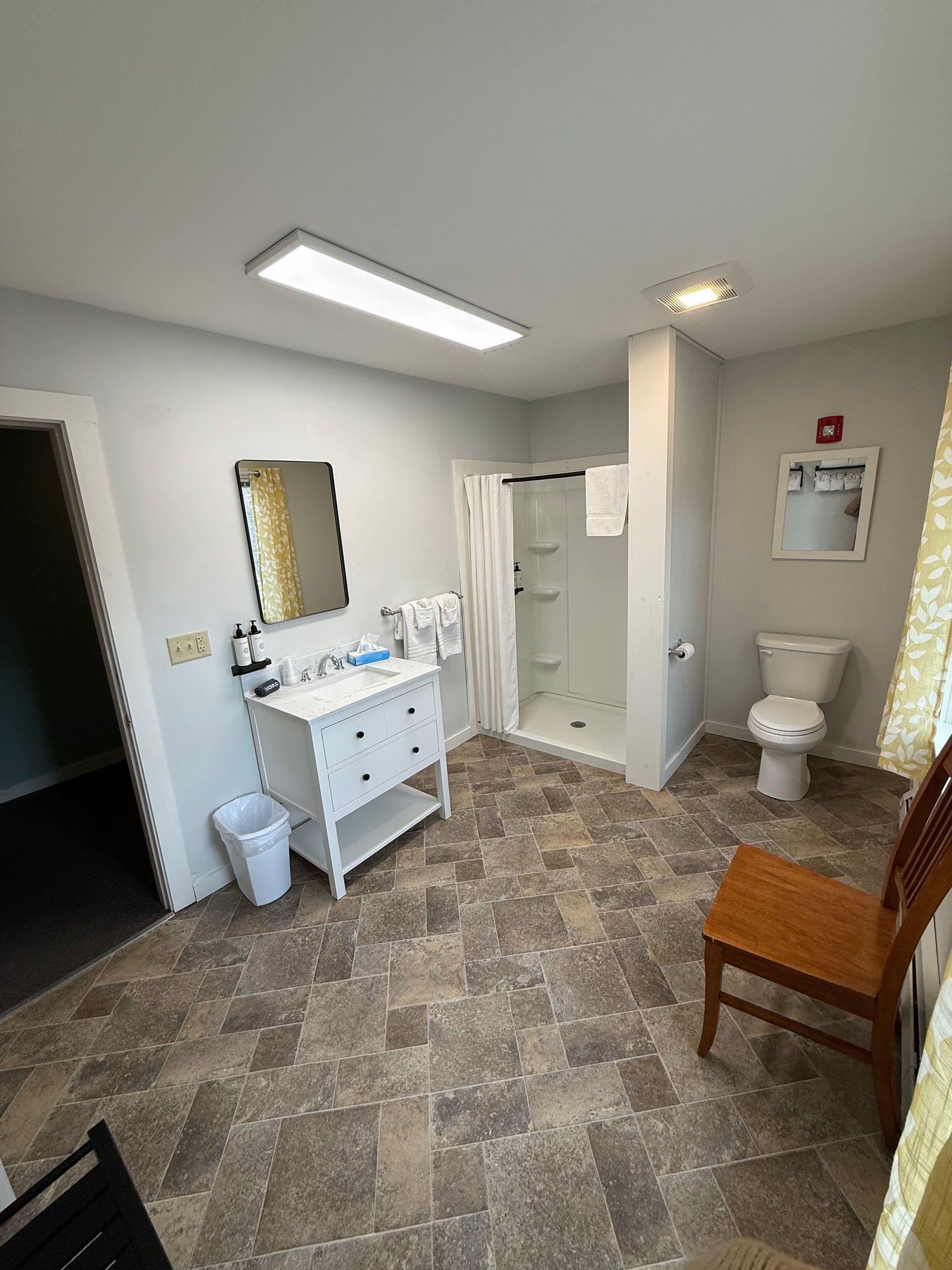 Small bathroom with vanity, shower, toilet, and wooden chair on patterned tile floor.