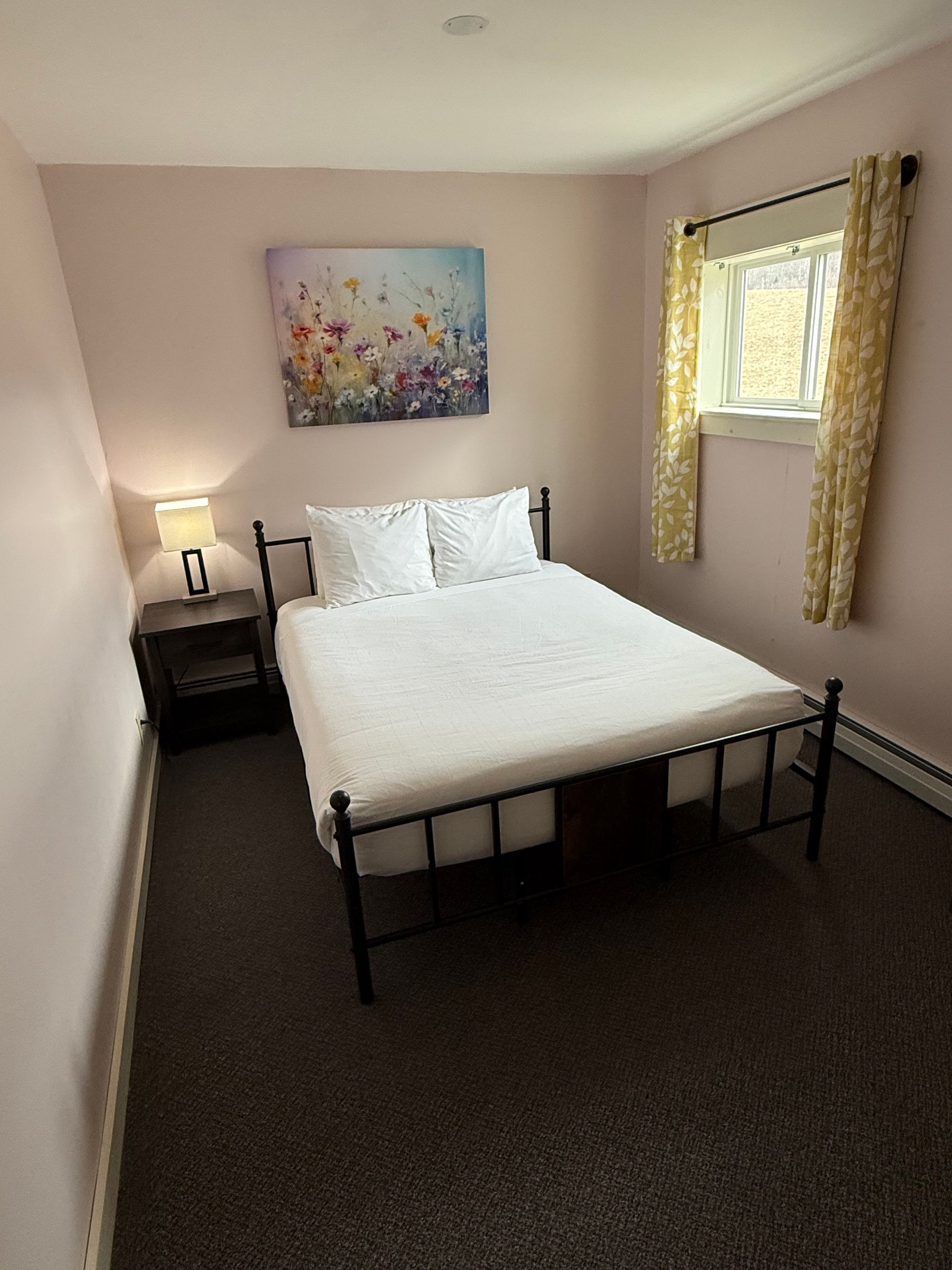 Small bedroom with a black bed frame, white bedding, bedside table, wall art, and a window with yellow curtains.