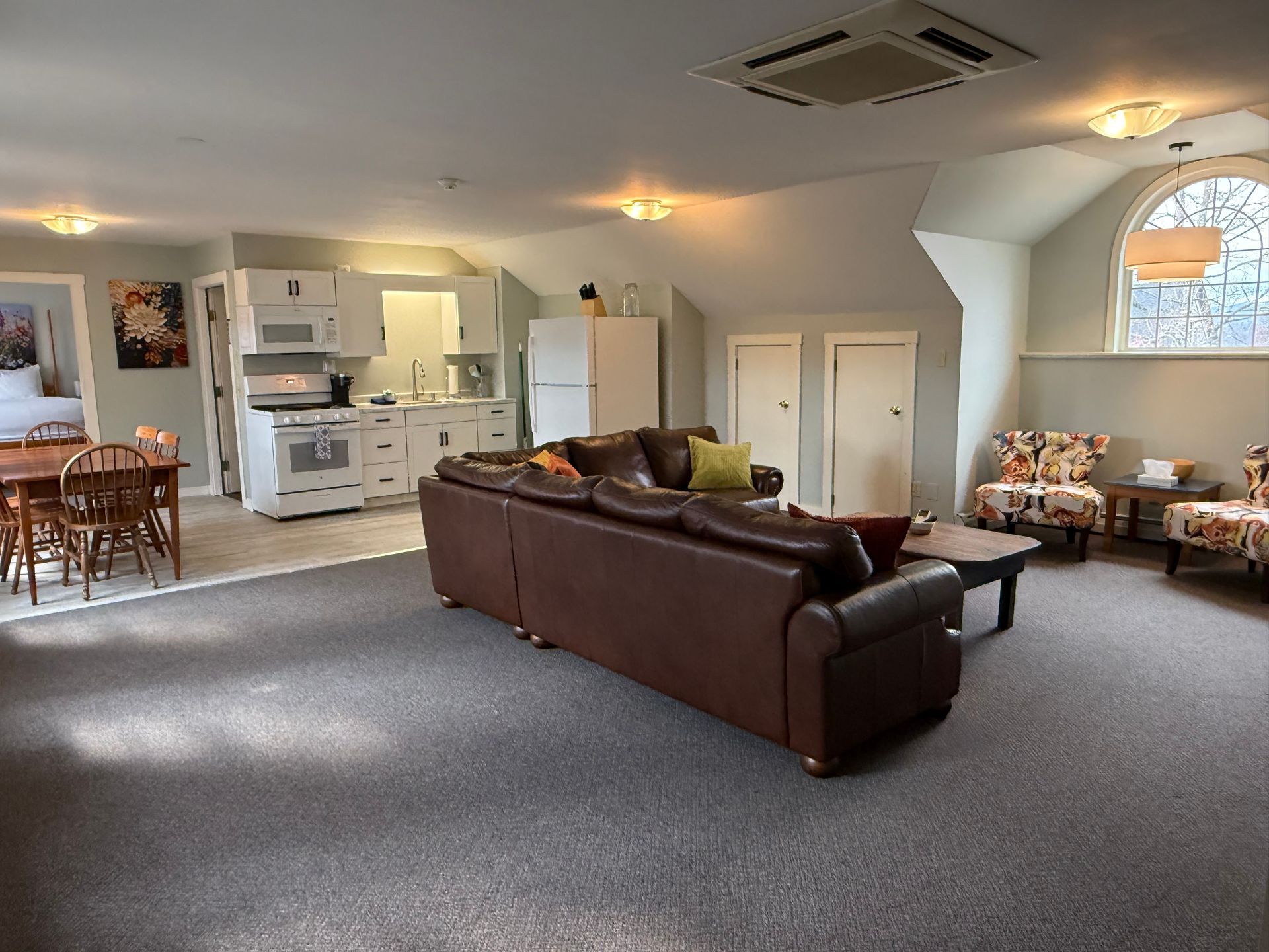 Spacious living room with brown sectional sofa, kitchen area, dining table, and arched window seating