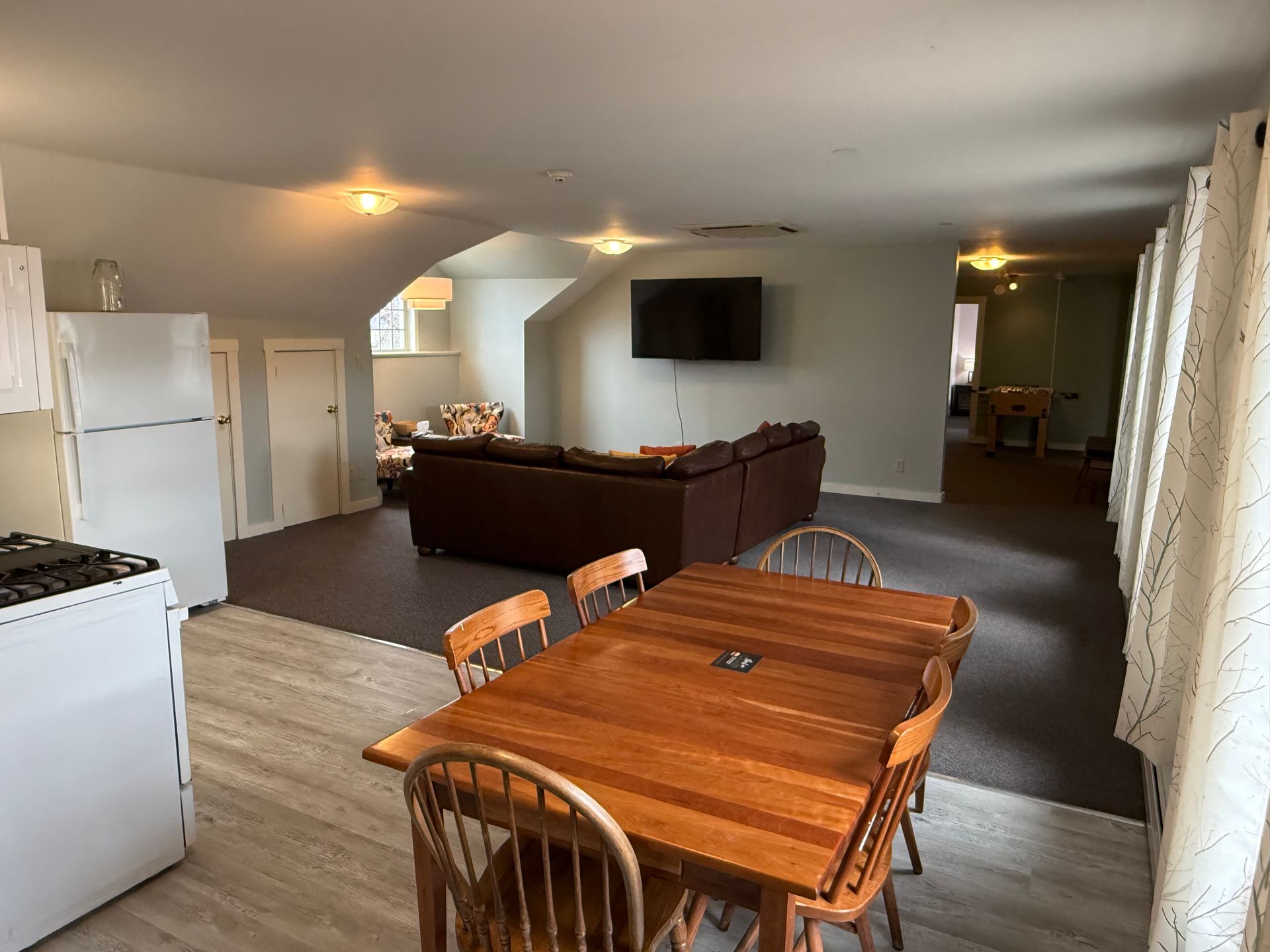Open-plan living room and dining area with wooden table, sectional sofa, TV, and kitchen appliances
