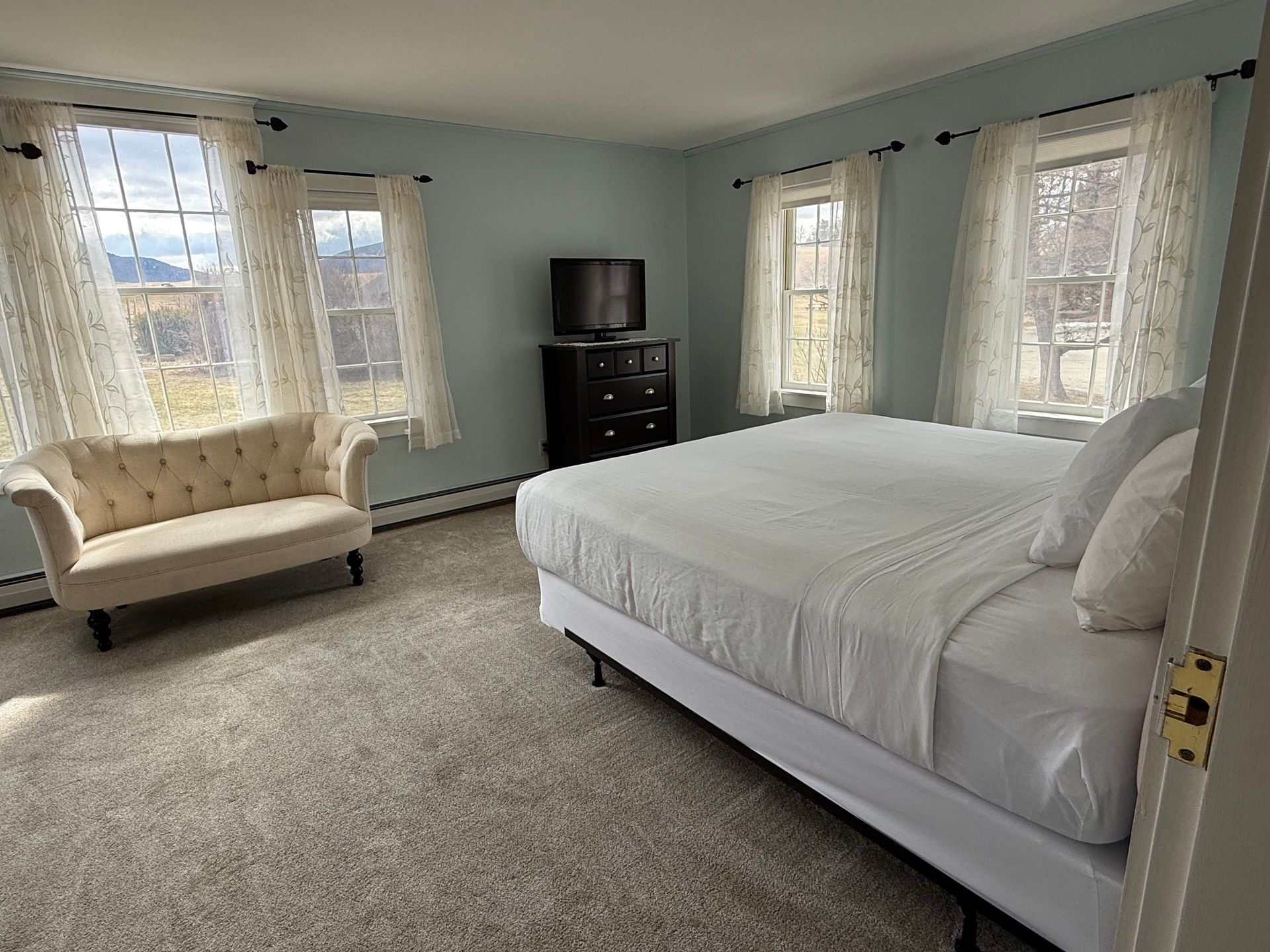 Bright bedroom with white bed, beige loveseat, black dresser, and lace-curtained windows