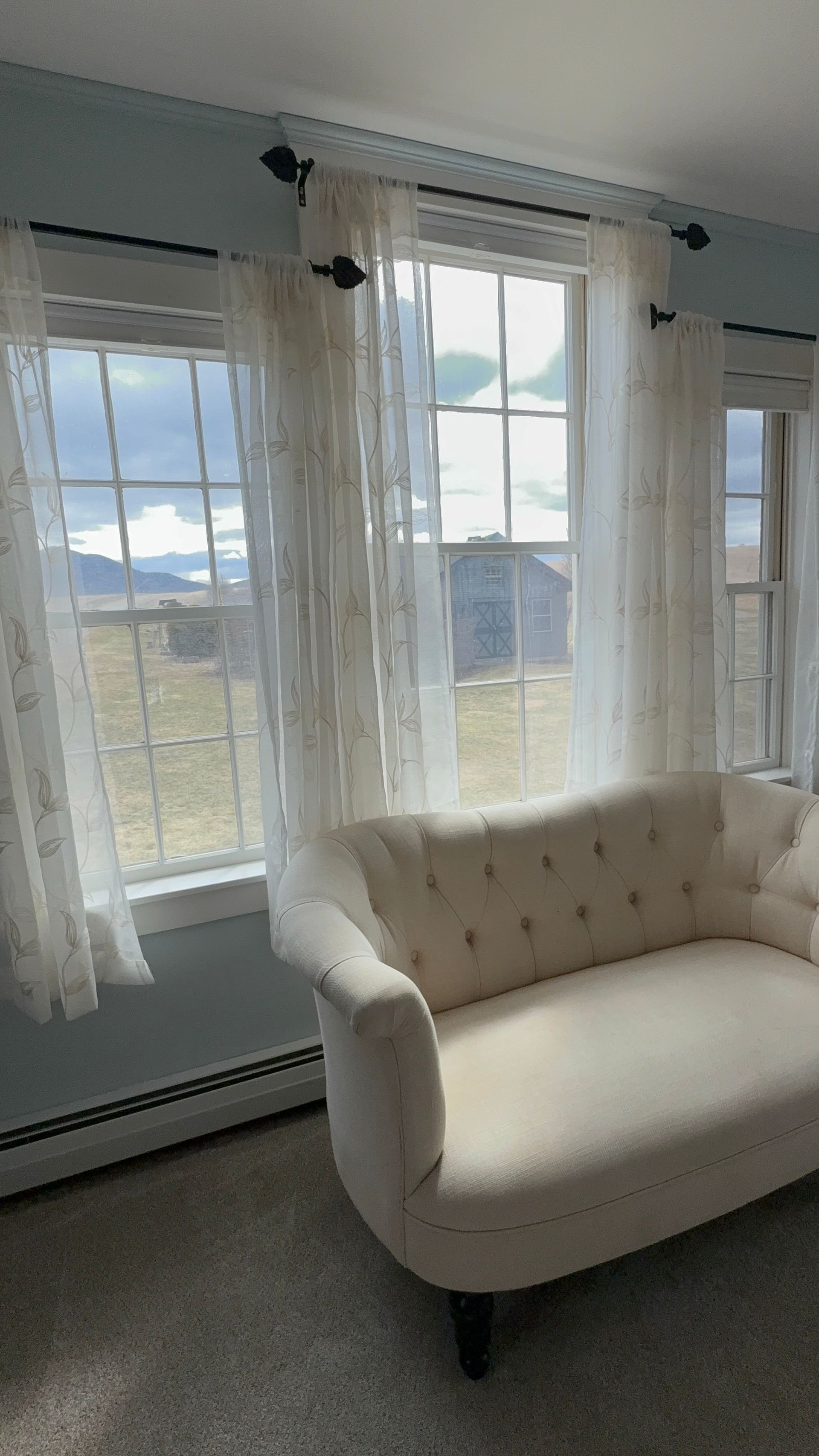 White tufted loveseat by large windows with sheer curtains and a mountain view.