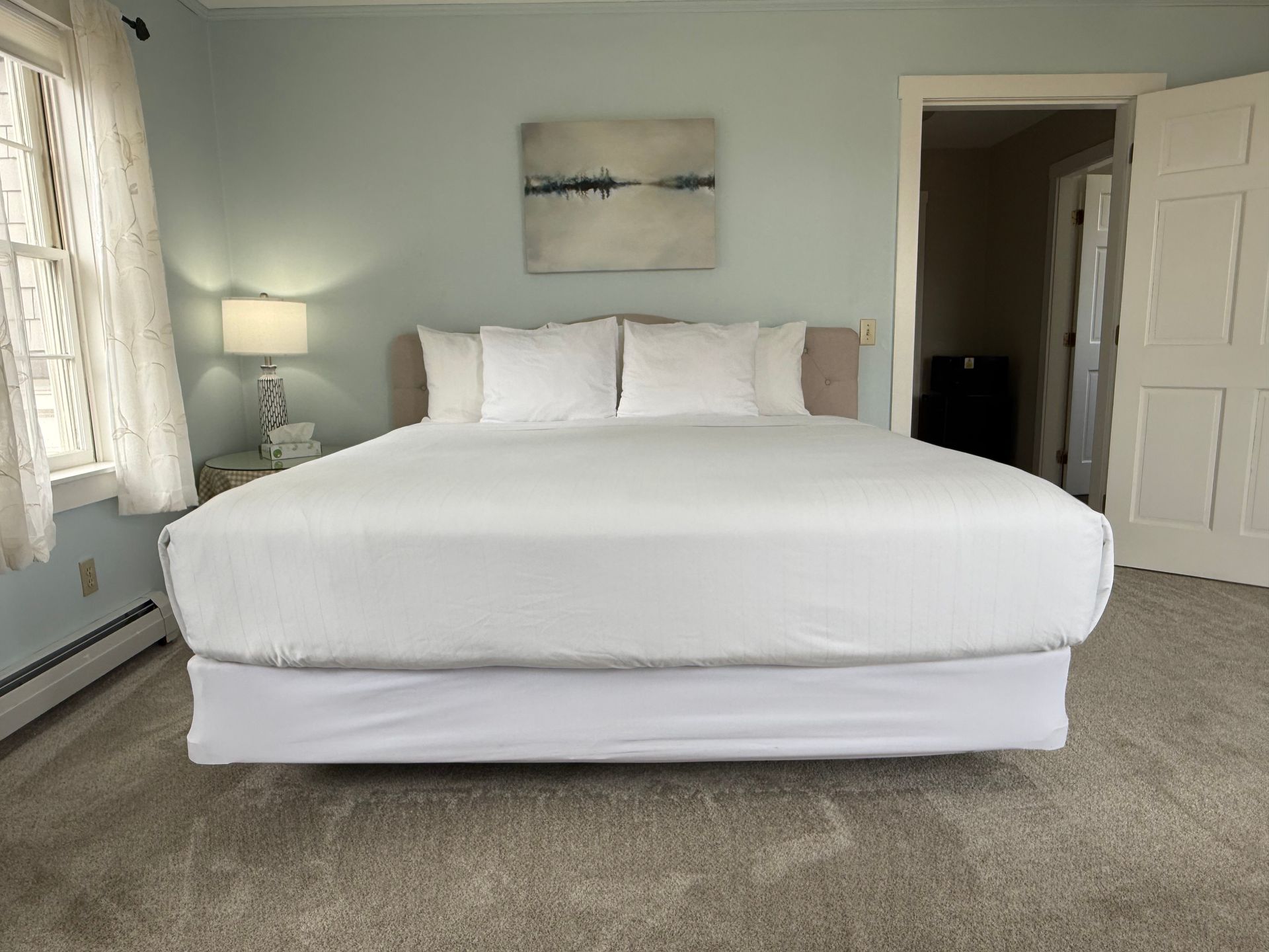 Bright bedroom with a white bed, lamp by window, and open door to hallway.