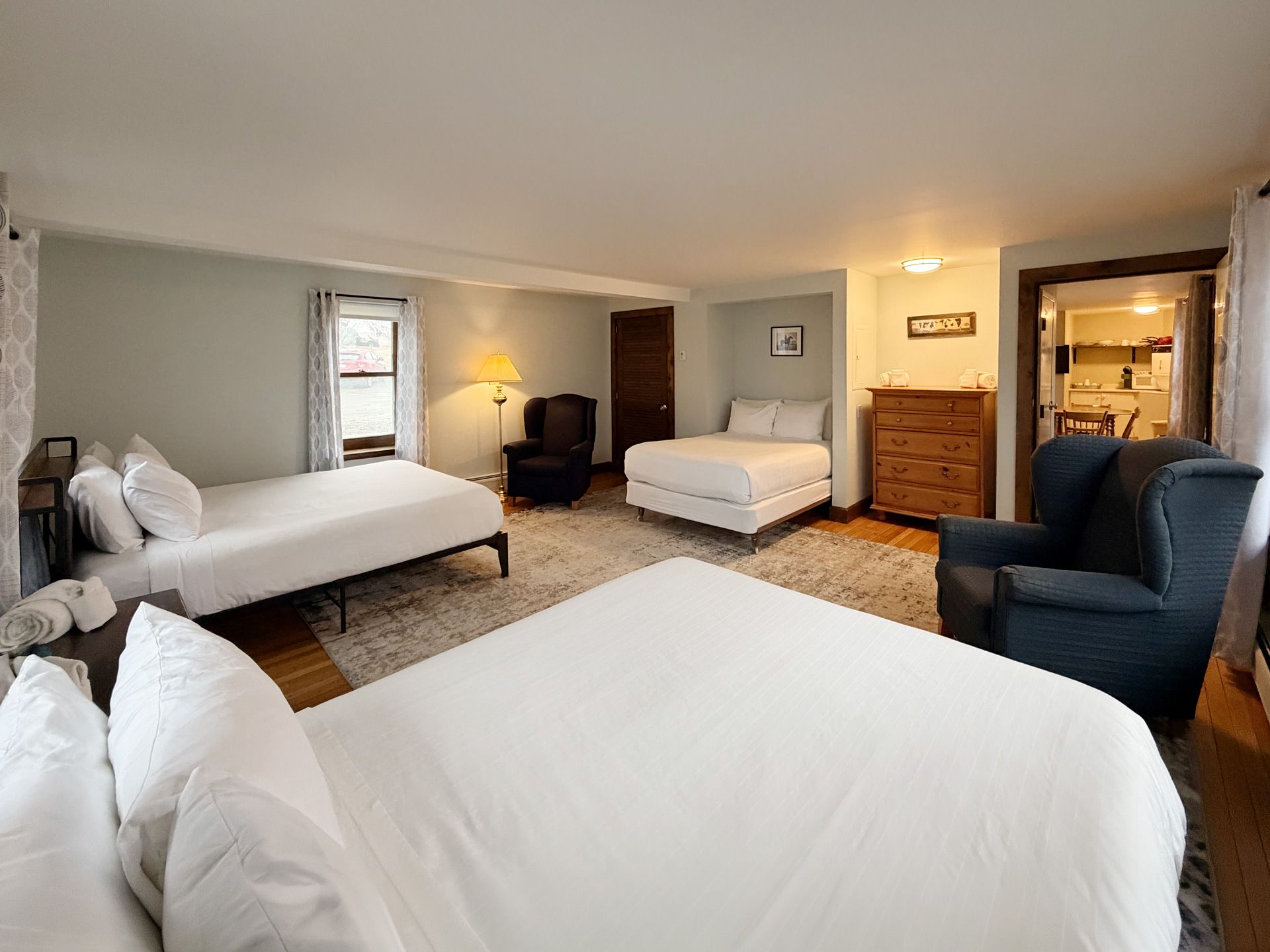 Spacious hotel room with four white beds, a blue armchair, dresser, and warm lighting