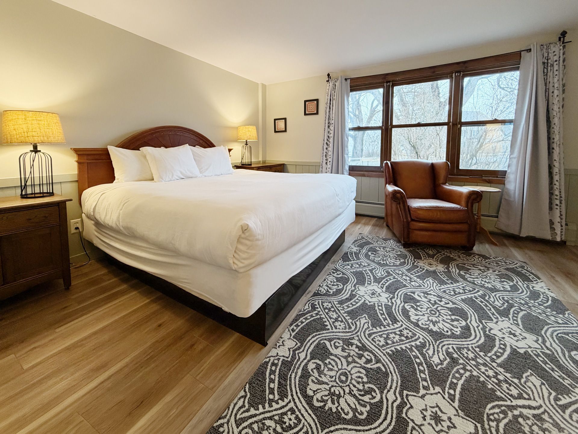 Cozy bedroom with a large bed, armchair by bay windows, and patterned rug on wood floors