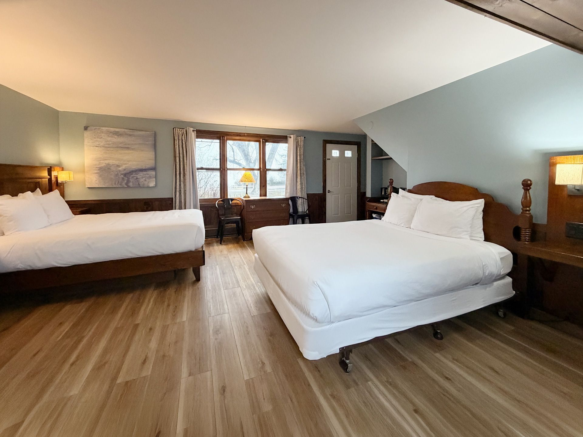 Spacious bedroom with two large white beds, wood floors, and a bright window centered on a cozy seating area