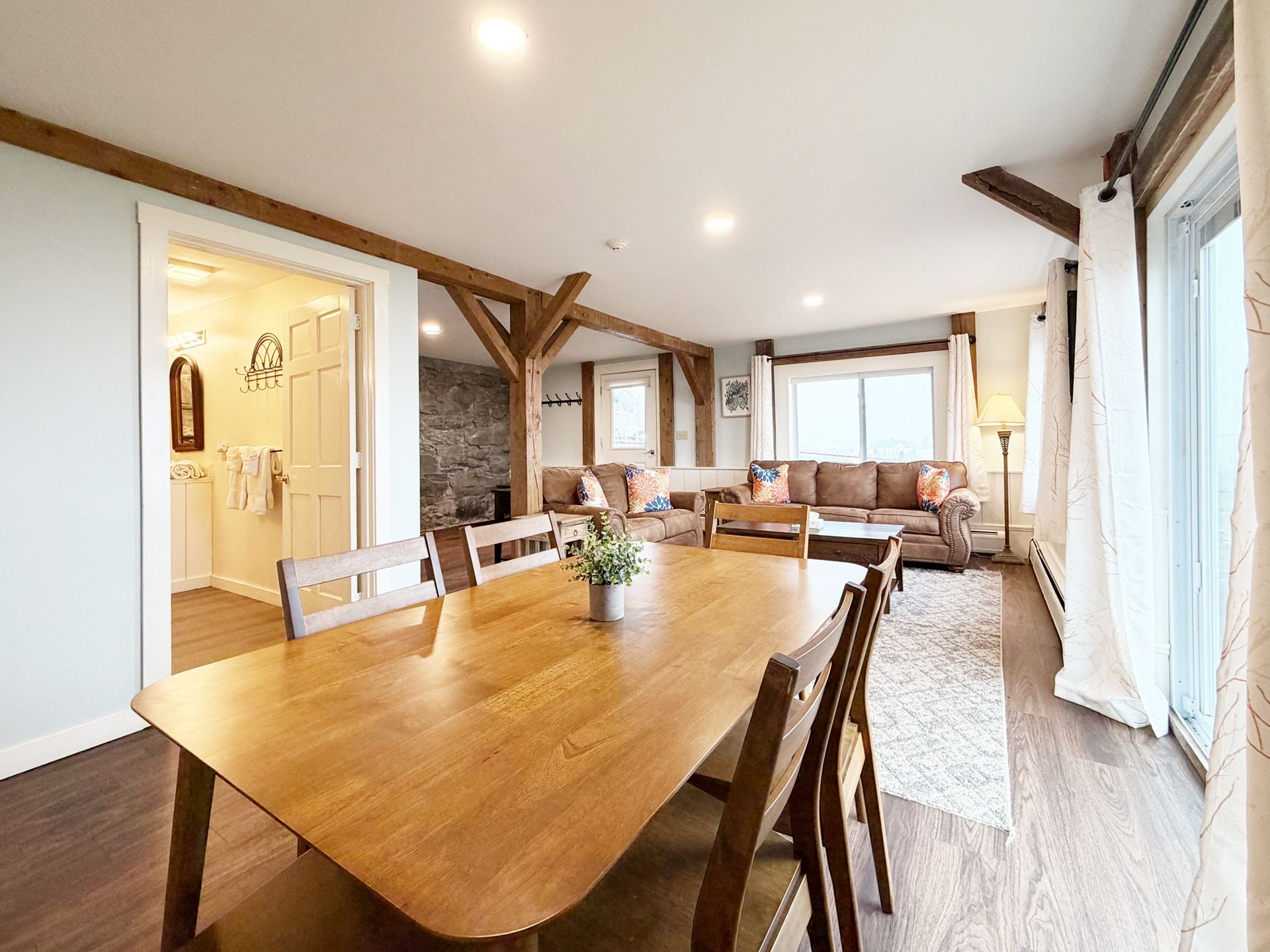 Bright dining and living room with wood beams, large table, sofa, and tall windows