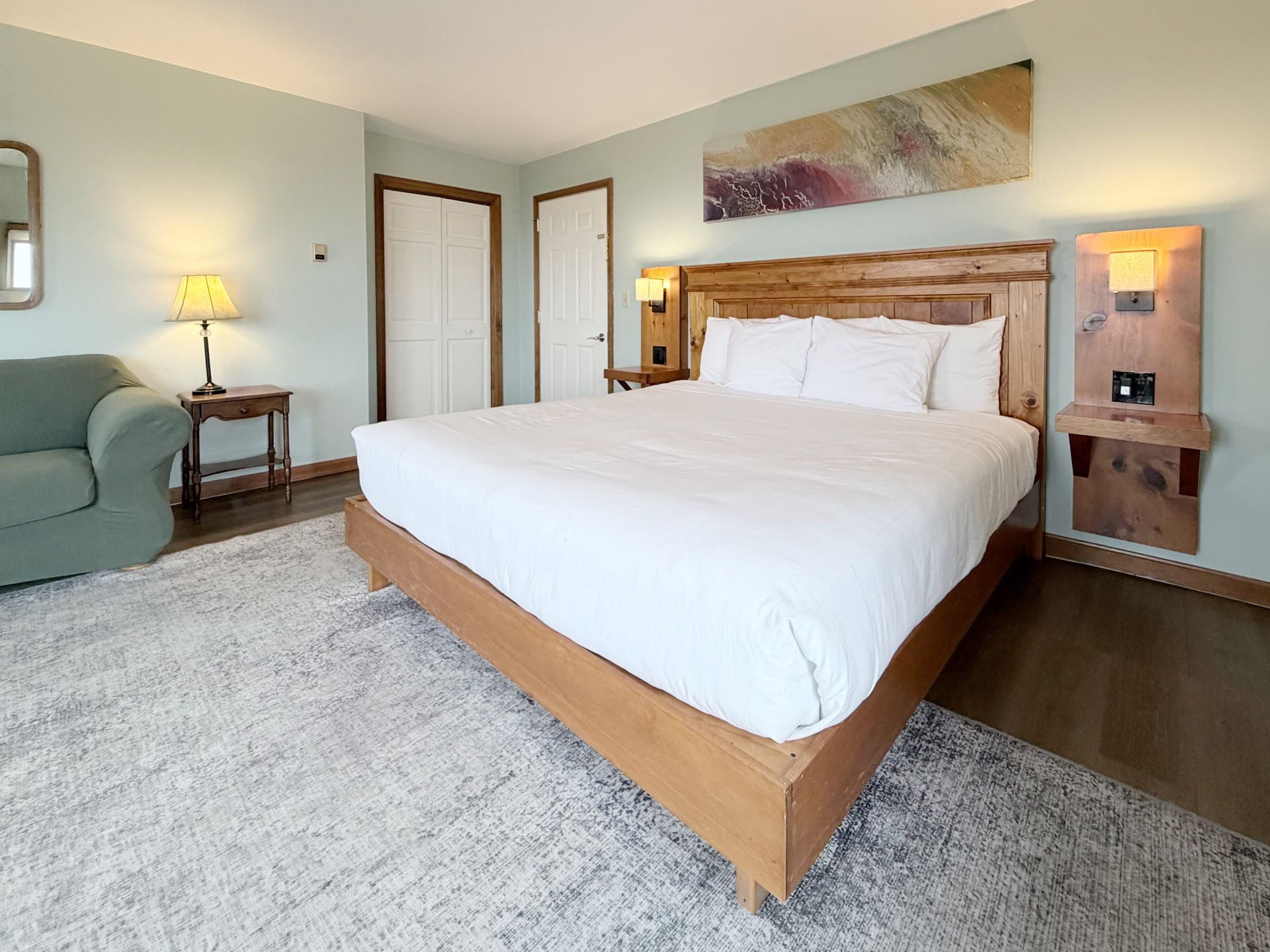 Bright, modern bedroom with a wooden bed, white bedding, gray rug, sofa, and bedside lamps.