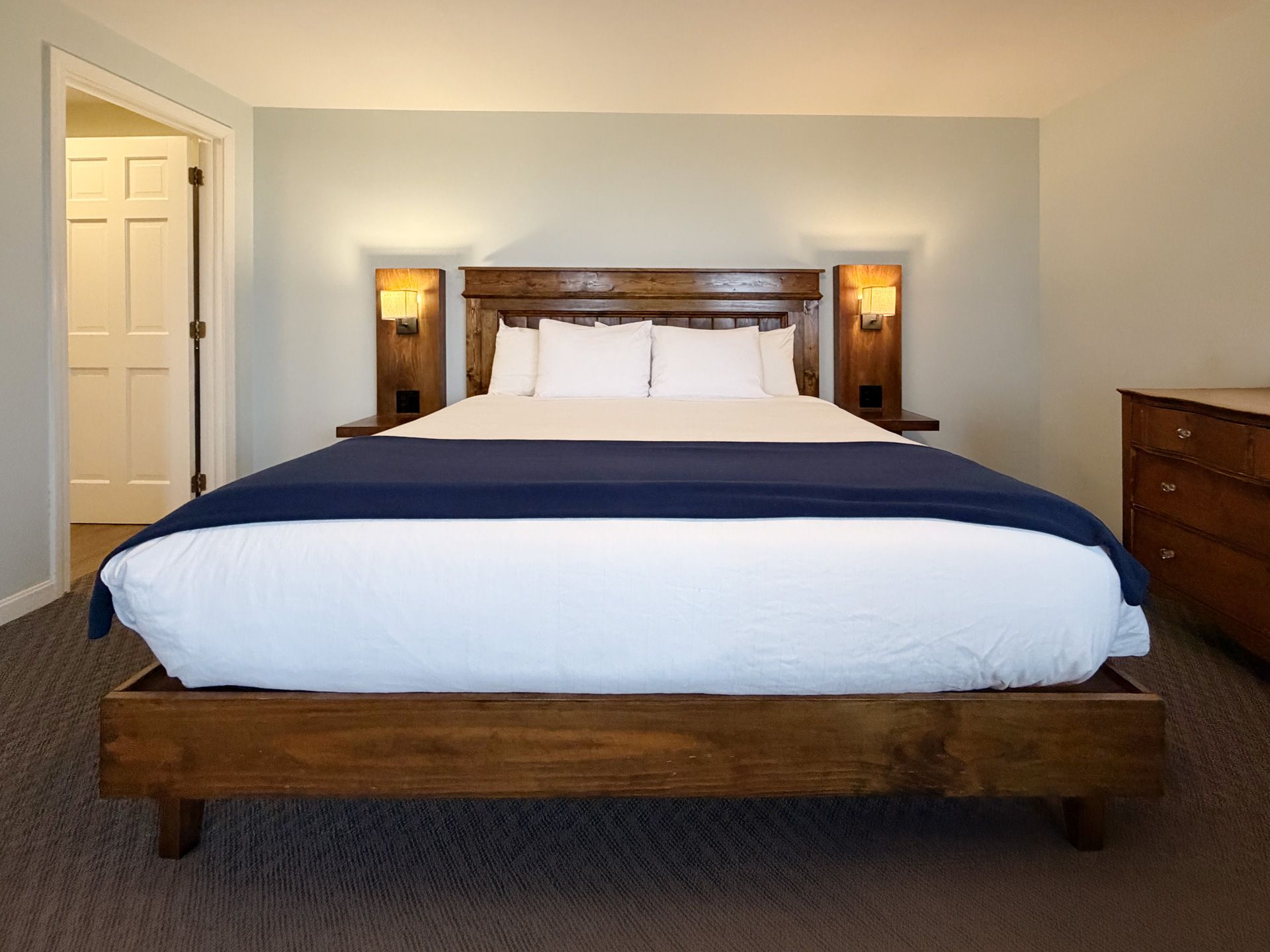 A neatly made bed with blue bedding in a softly lit bedroom, with lamps on each side and a dresser.