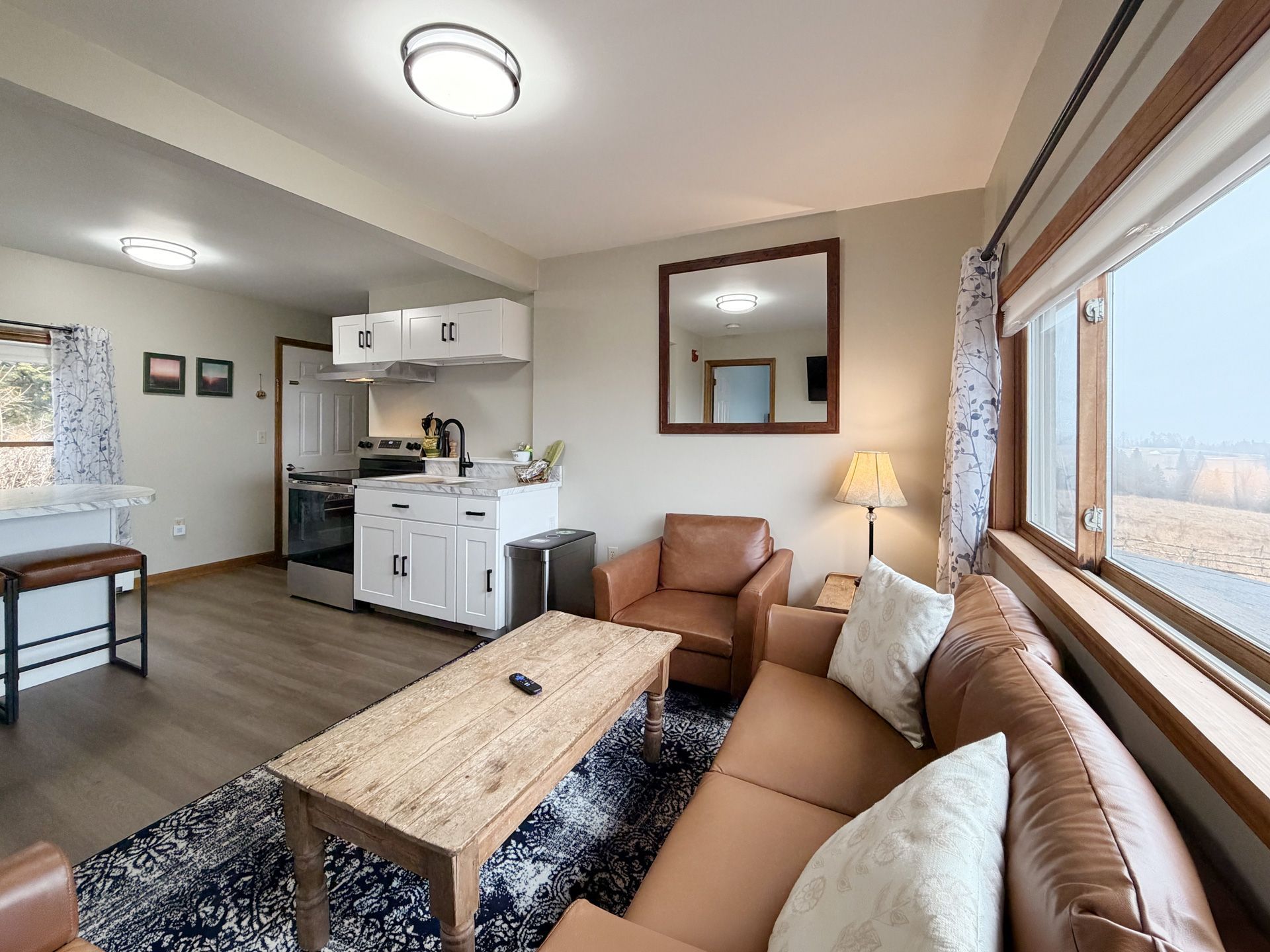 Bright living room with tan sectional, coffee table, patterned rug, and open kitchen area by large windows.