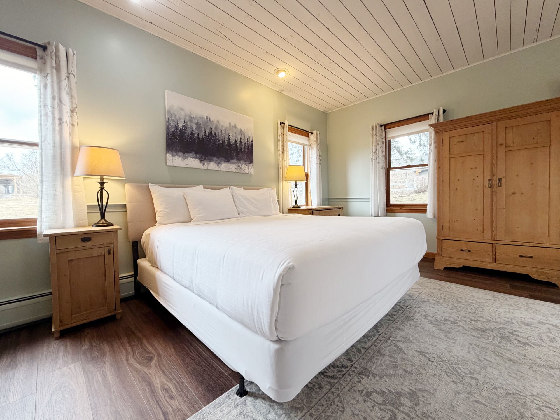 Bright bedroom with a white bed, wooden wardrobe, lamp, and large windows in a cozy neutral-toned room