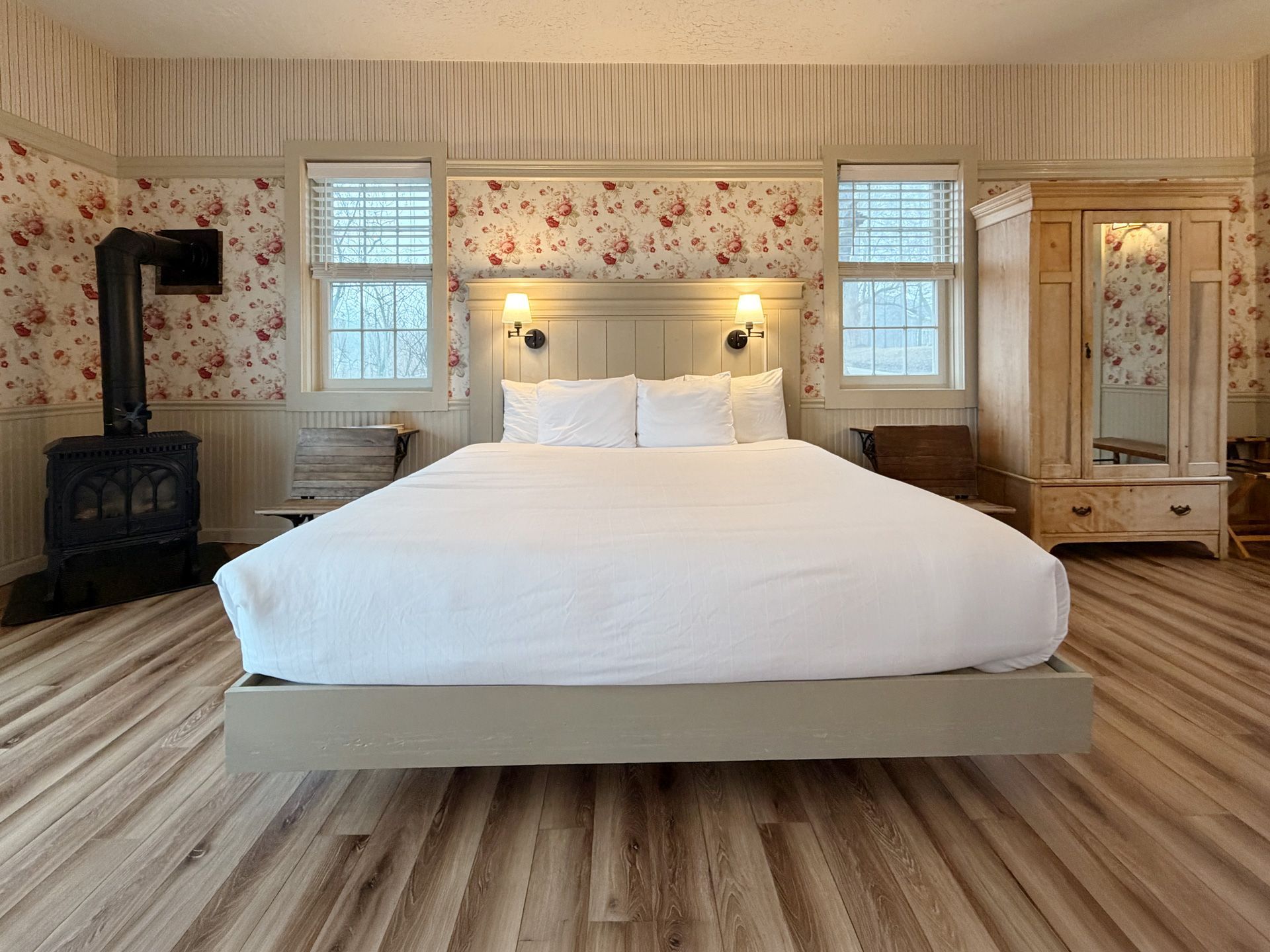 Bright bedroom with a large white bed, floral wallpaper, wood floors, and a black wood stove.