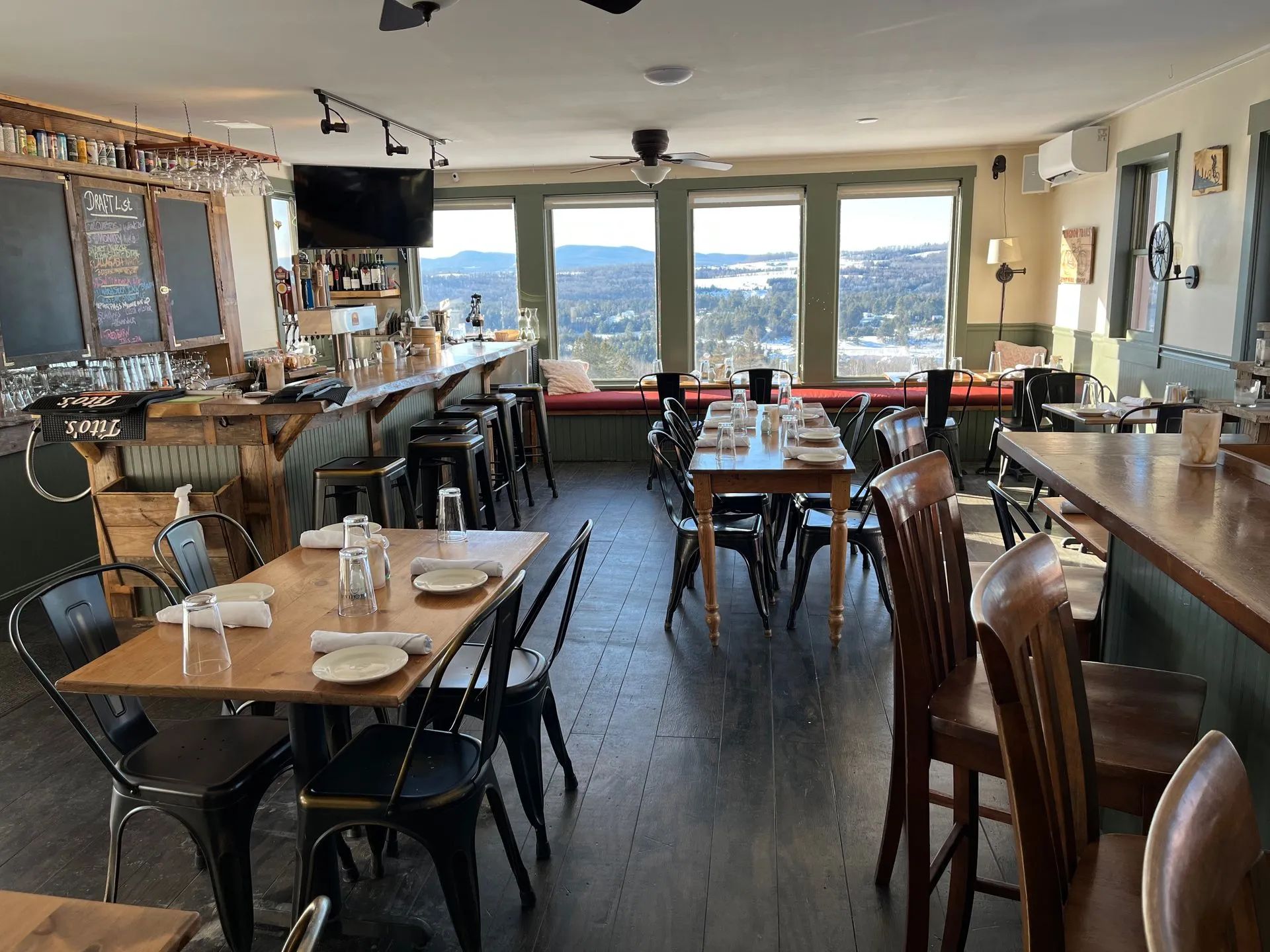 Empty restaurant dining area with tables, chairs, bar, and large windows overlooking a city view.