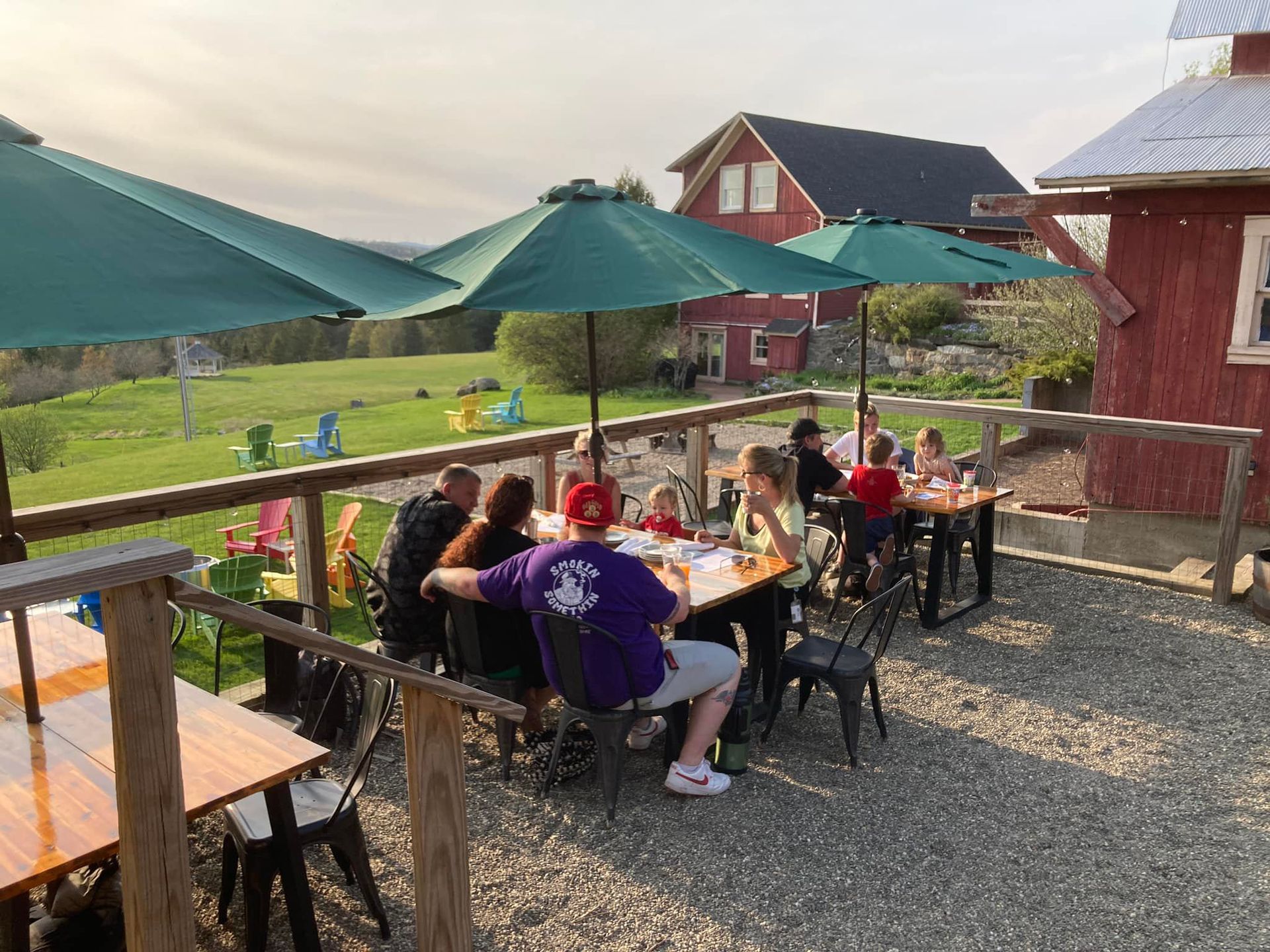 People seated at outdoor tables under green umbrellas on a patio beside rustic red buildings.