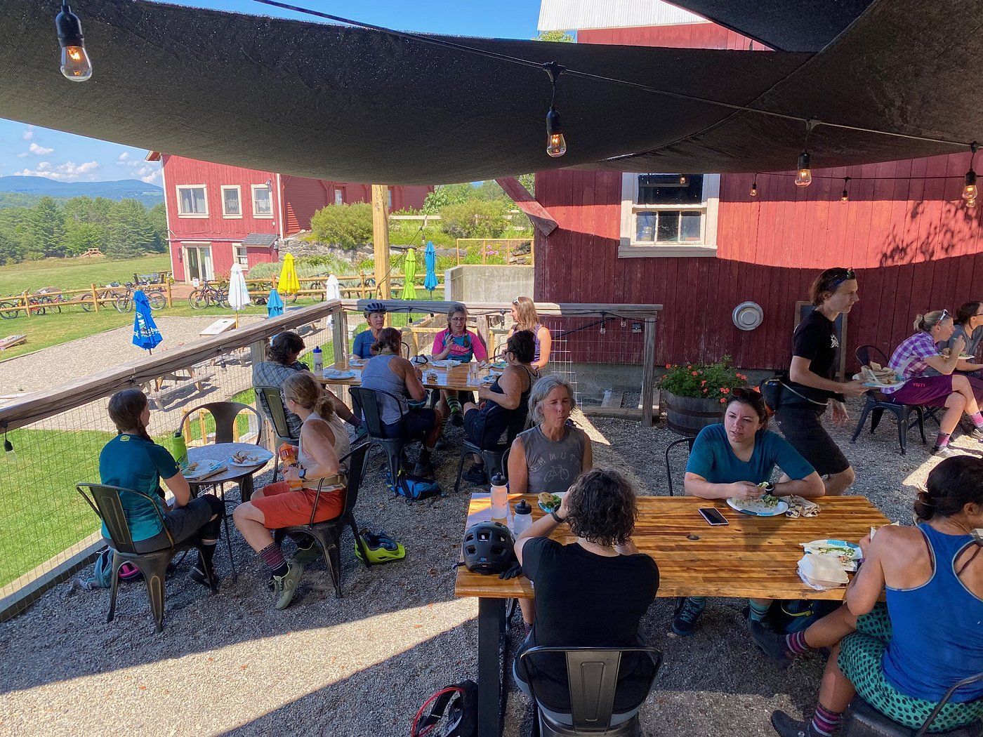People seated at picnic tables under a shade canopy beside a red barn on a sunny day.
