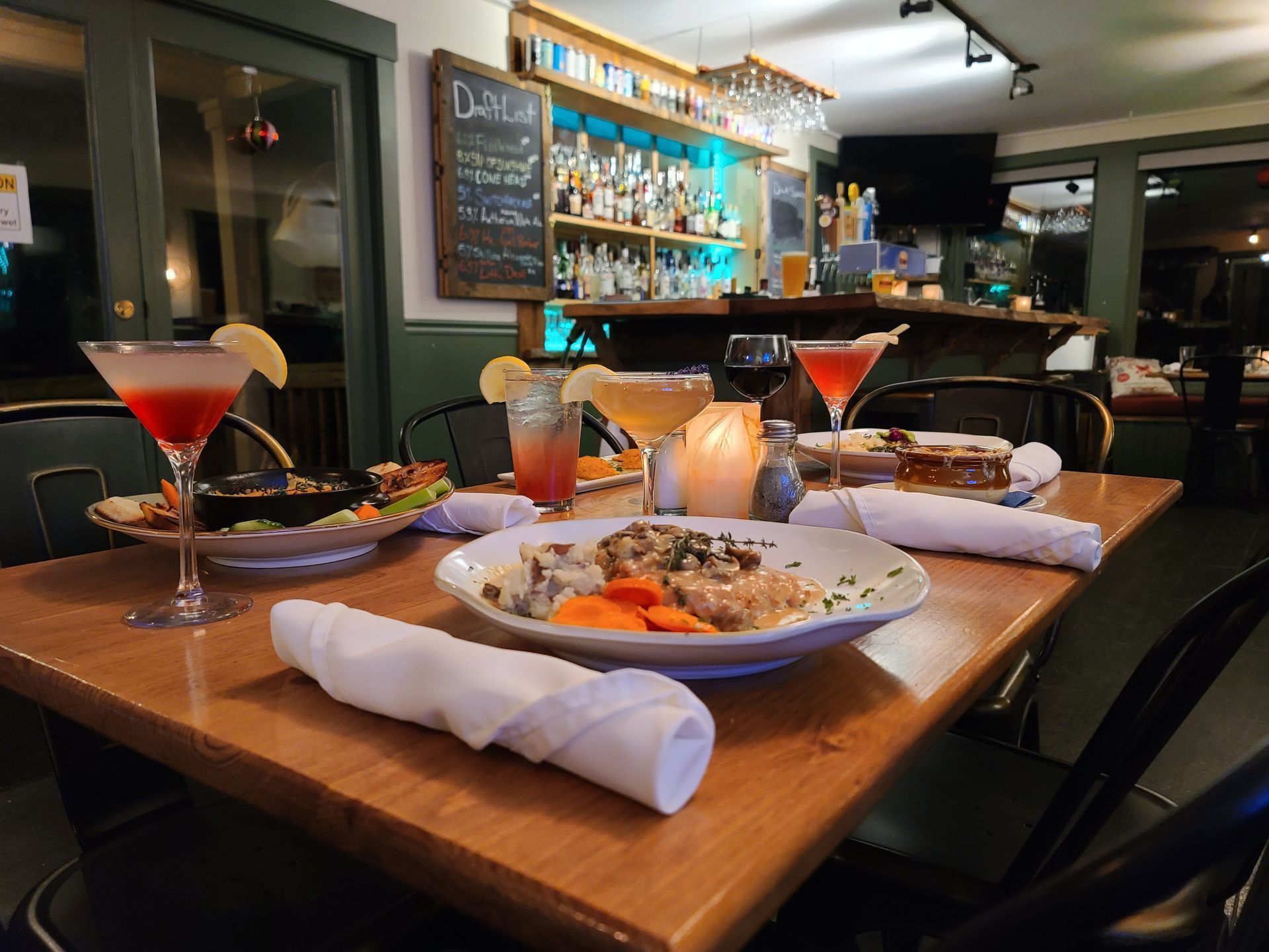 Restaurant table with cocktails, plated meals, and a lit bar in the background
