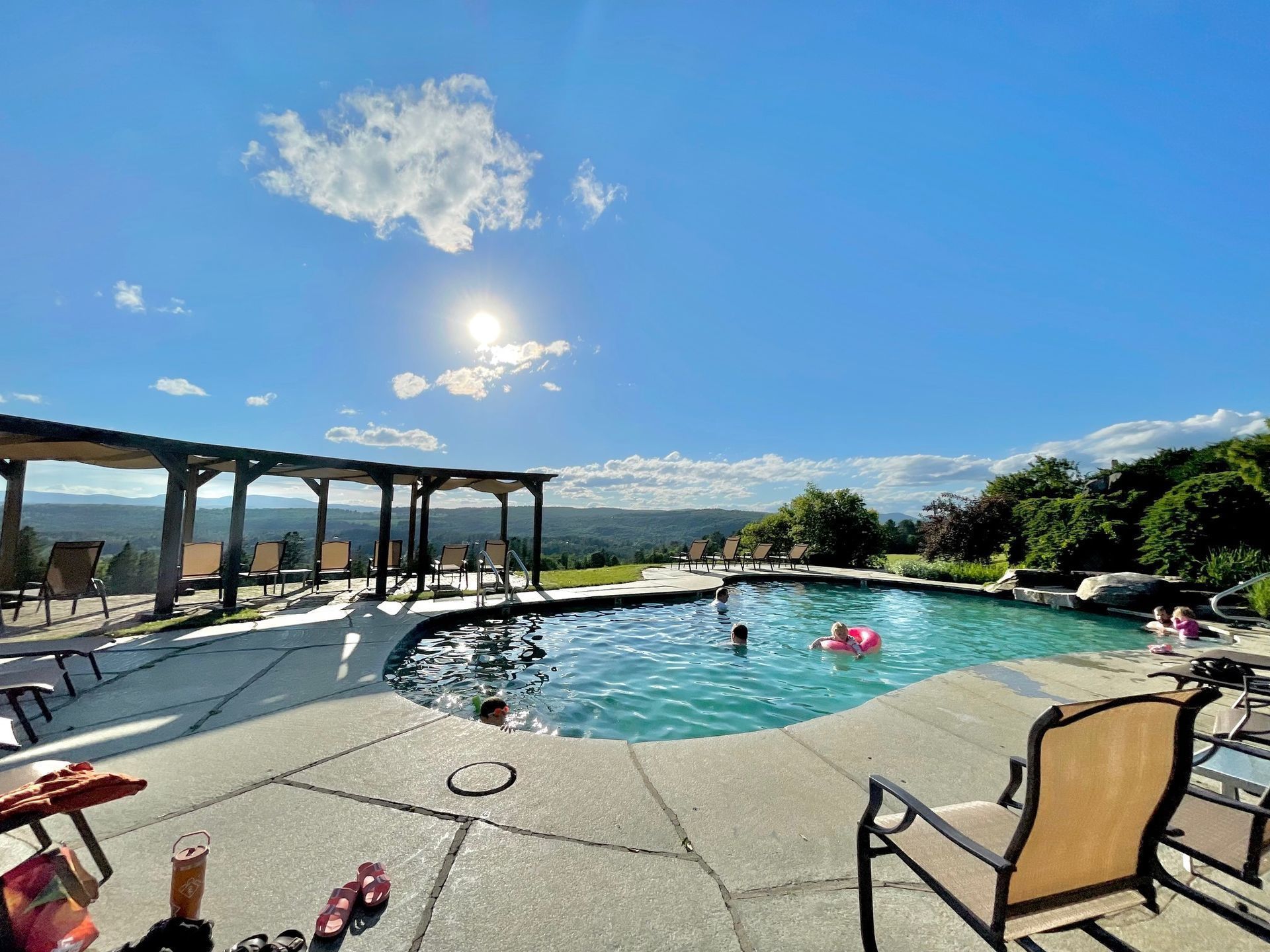 Poolside view of a resort pool with lounge chairs, pergola shade, and bright blue sky over mountains