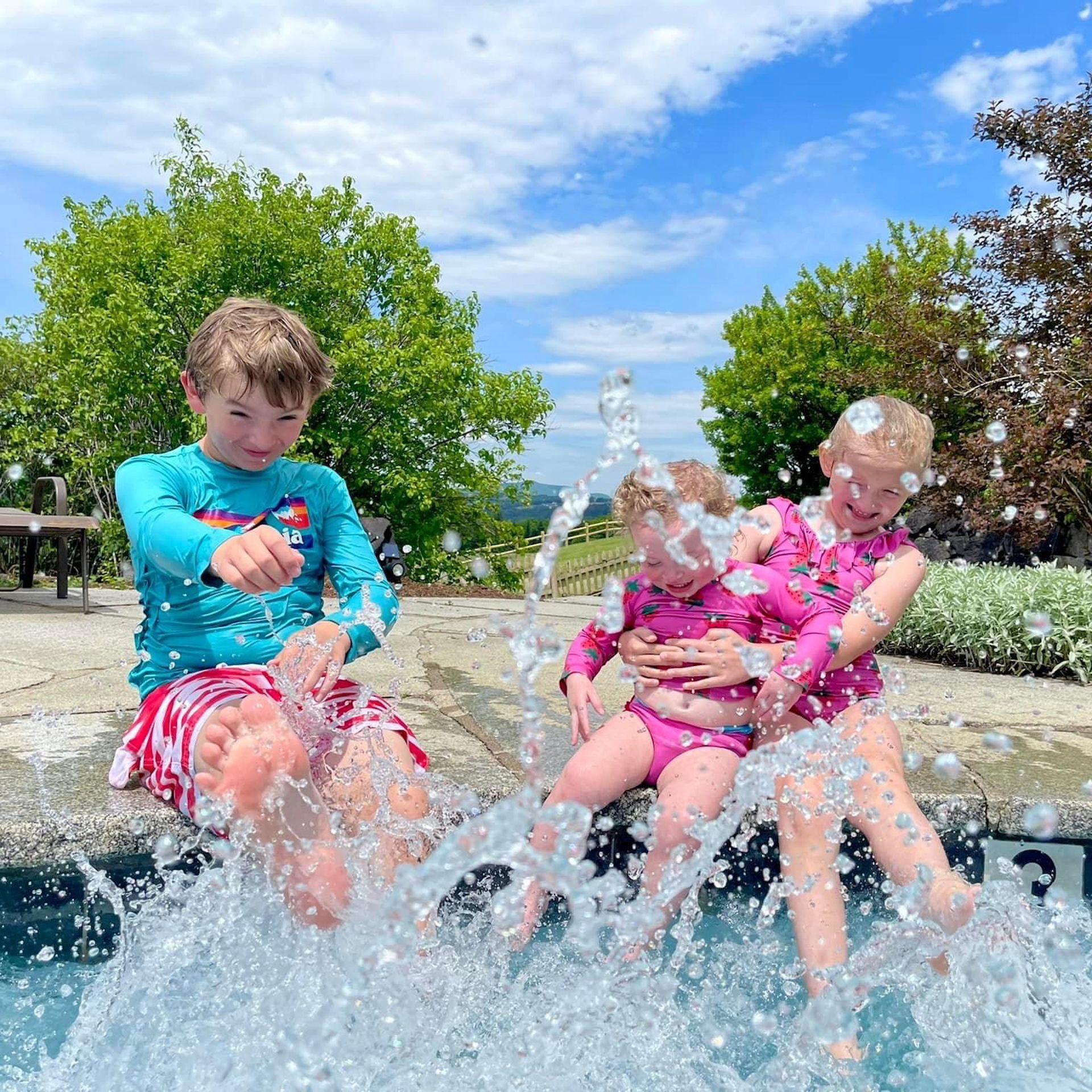 Three children splashing in a fountain on a sunny day, with water spraying around them.
