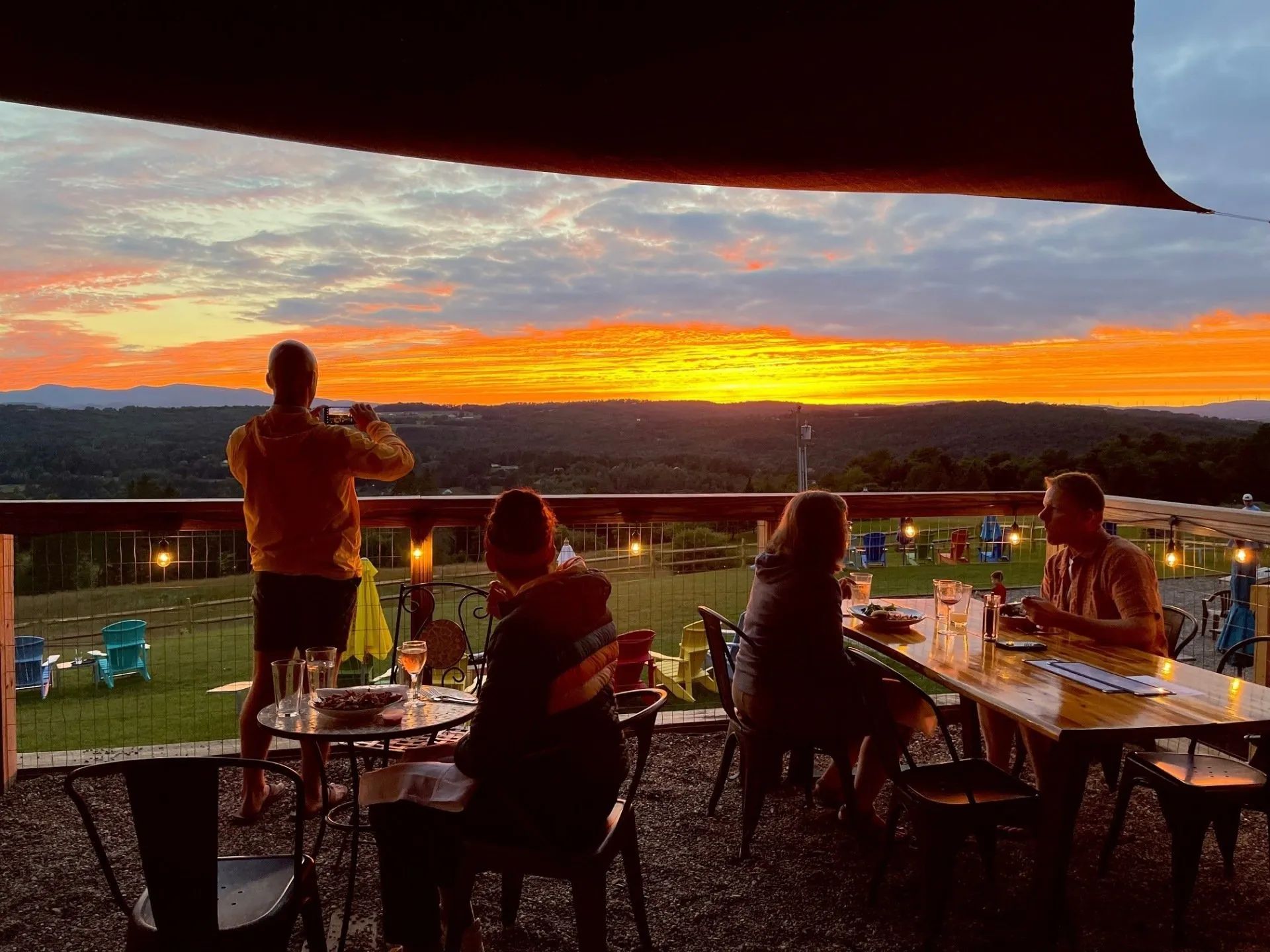 People dining on a patio at sunset, with a glowing orange sky over the landscape.