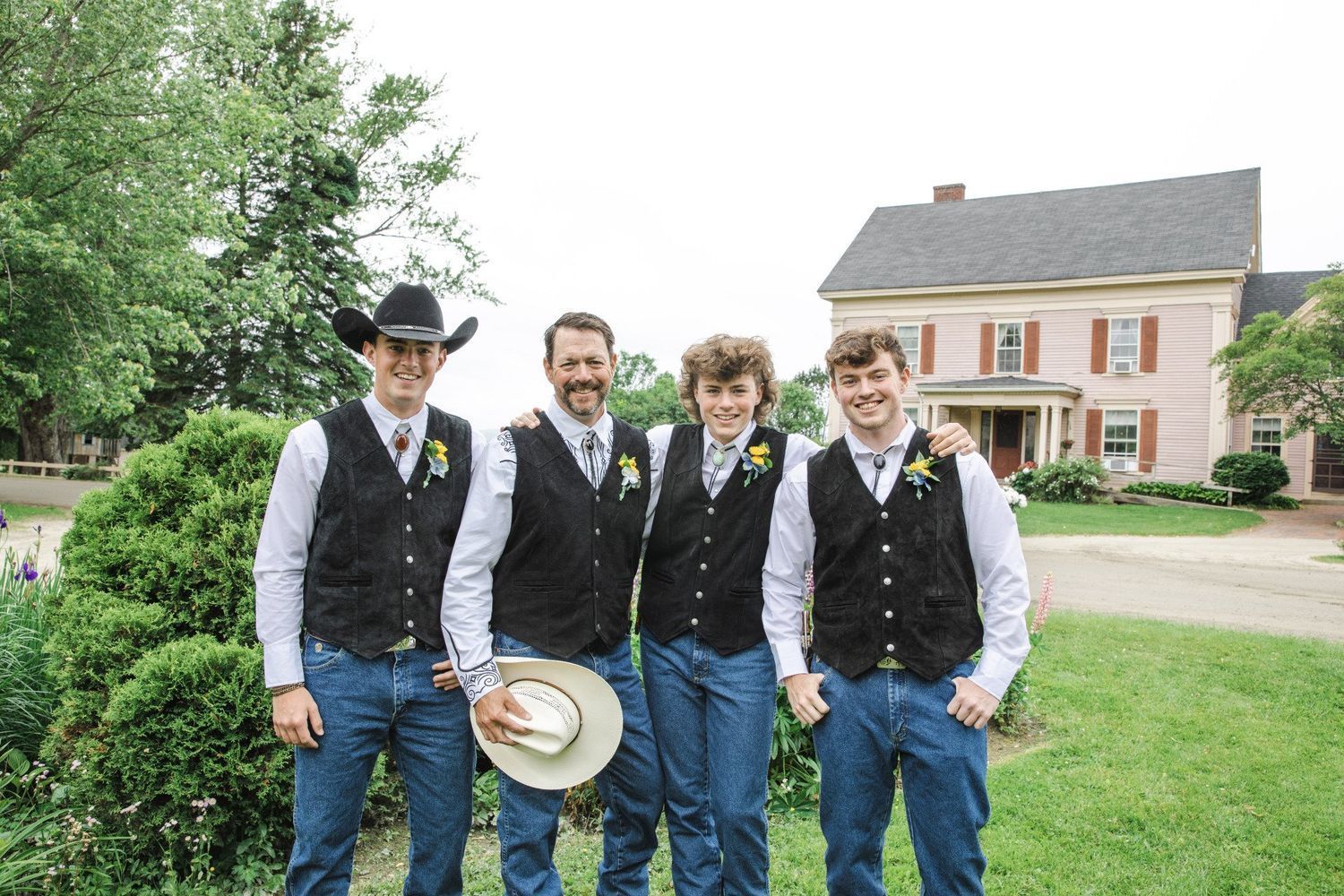 Four men in matching vests and jeans posing outdoors near a large house, one holding a cowboy hat