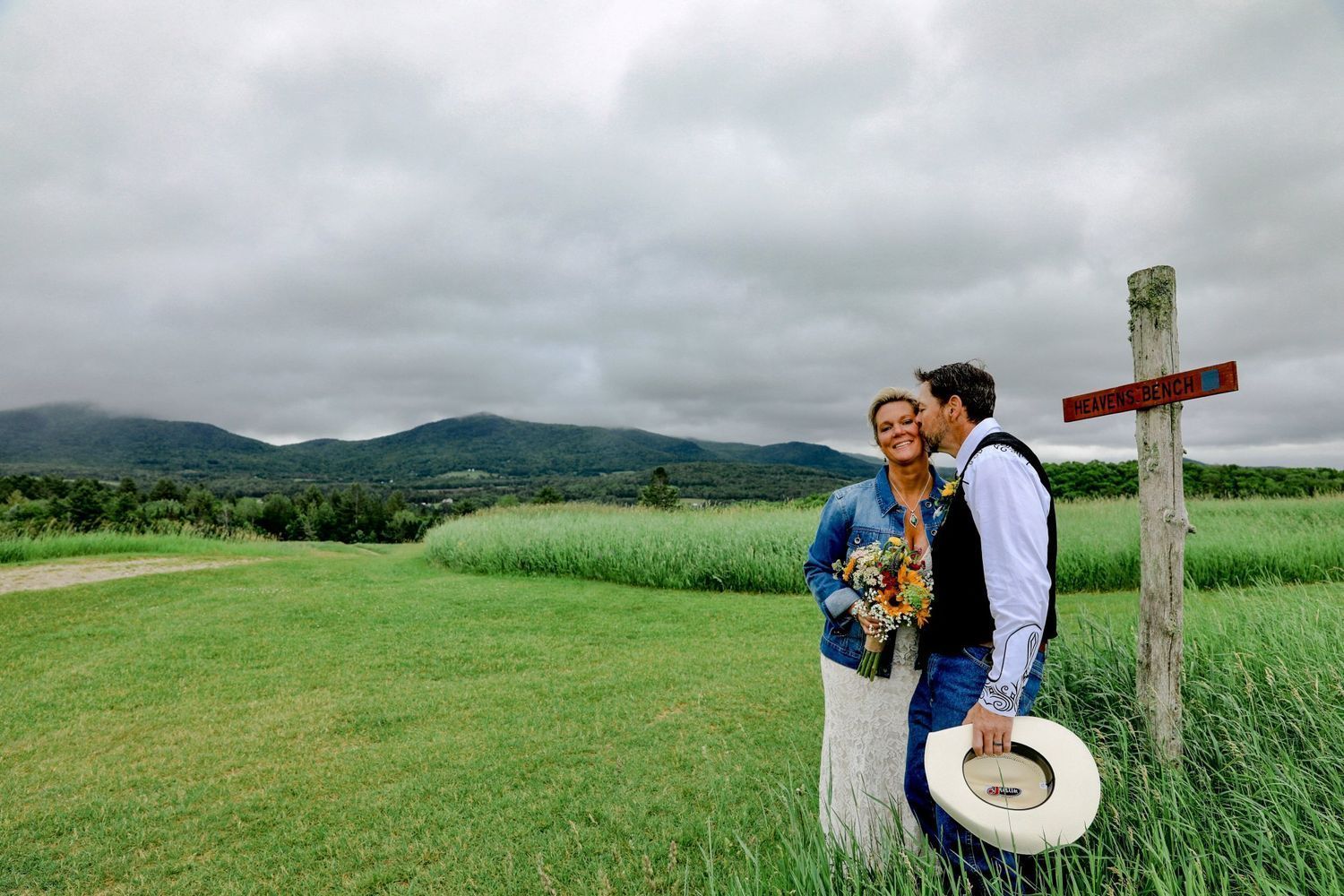 Couple standing in a green field by a wooden cross, with hills and cloudy sky in the background.
