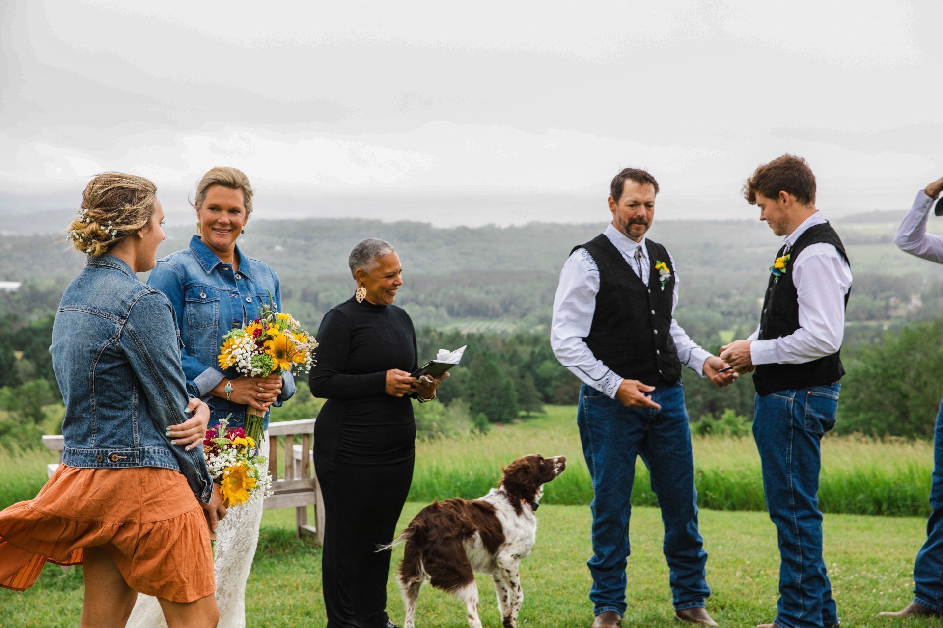 People standing with bouquets and a dog in a grassy field, with hills in the background.