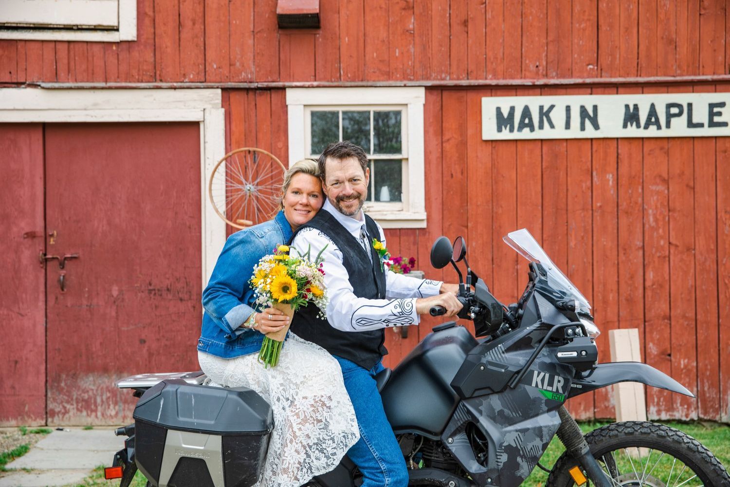 Smiling couple on a motorcycle in front of a red barn, with a bride holding sunflowers and veil