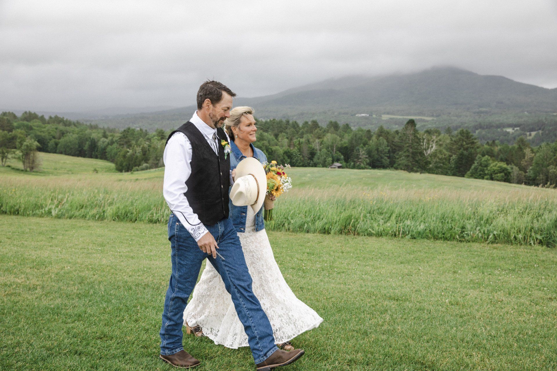 Couple walking through a grassy field, with mountains and cloudy sky in the background.