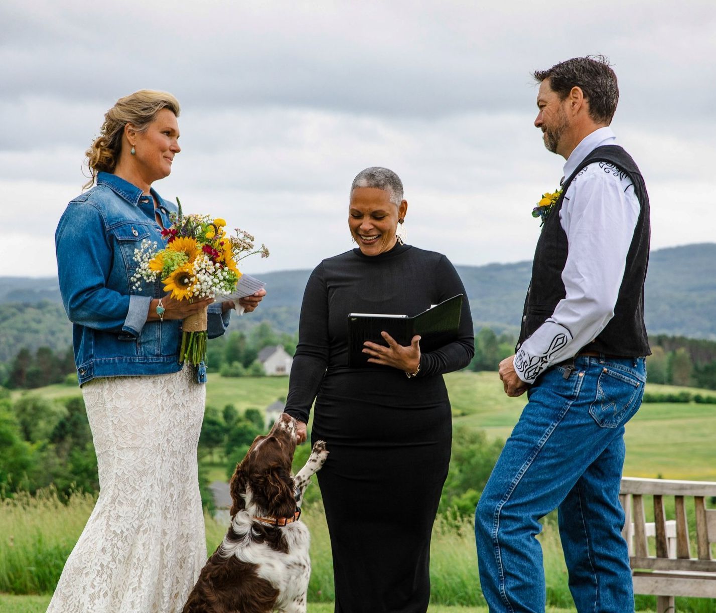 Three people and a dog stand outdoors during a small wedding ceremony, one holding flowers and one reading from a tablet.