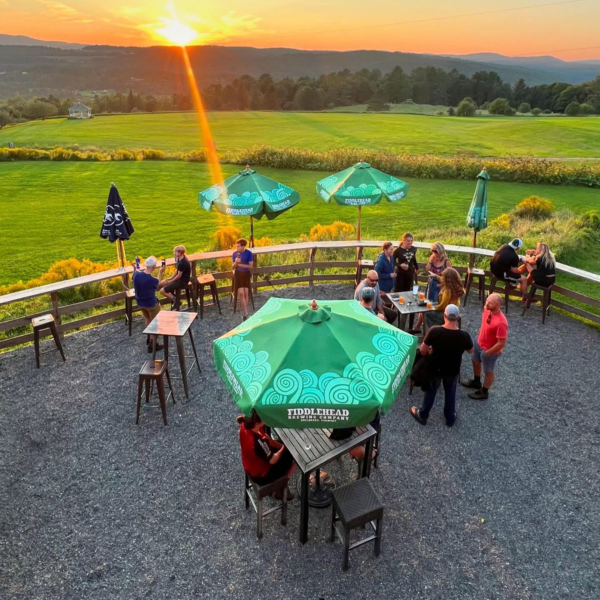 People on a patio with green umbrellas watching sunset over a field and distant hills.