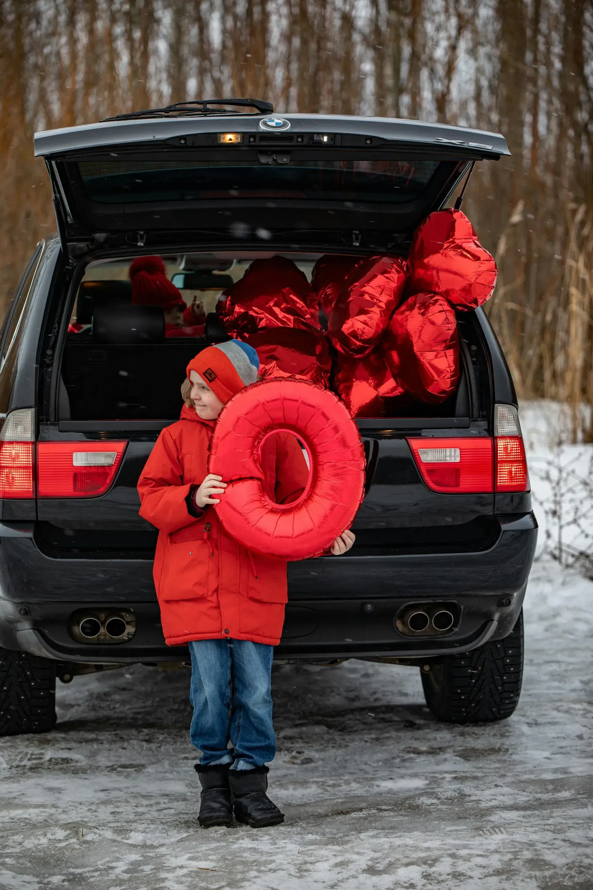 Child in red winter coat holding a red inflatable ring beside an open SUV in a snowy lot