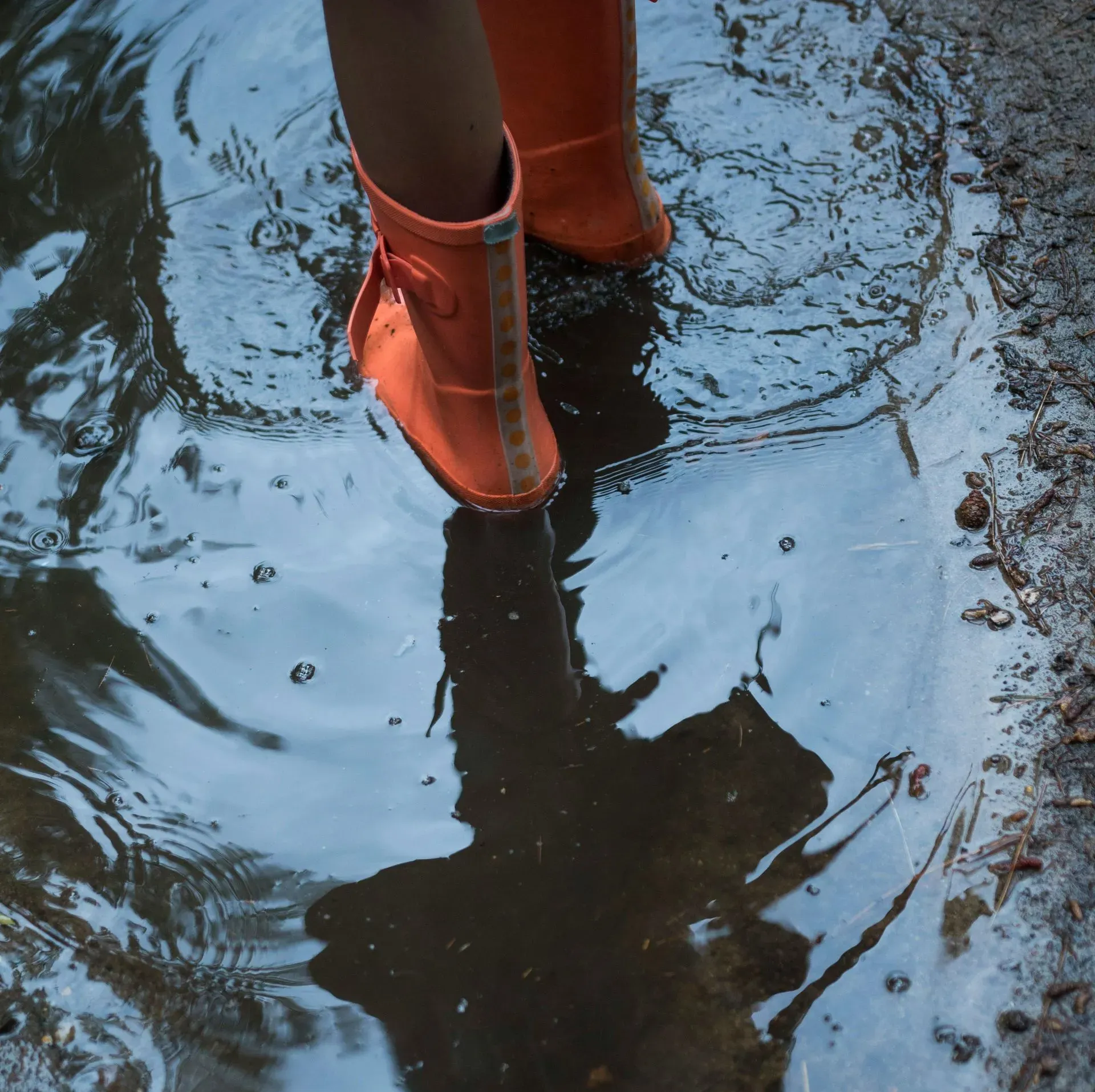 Orange rain boots standing in a puddle, with ripples reflecting the legs and sky.