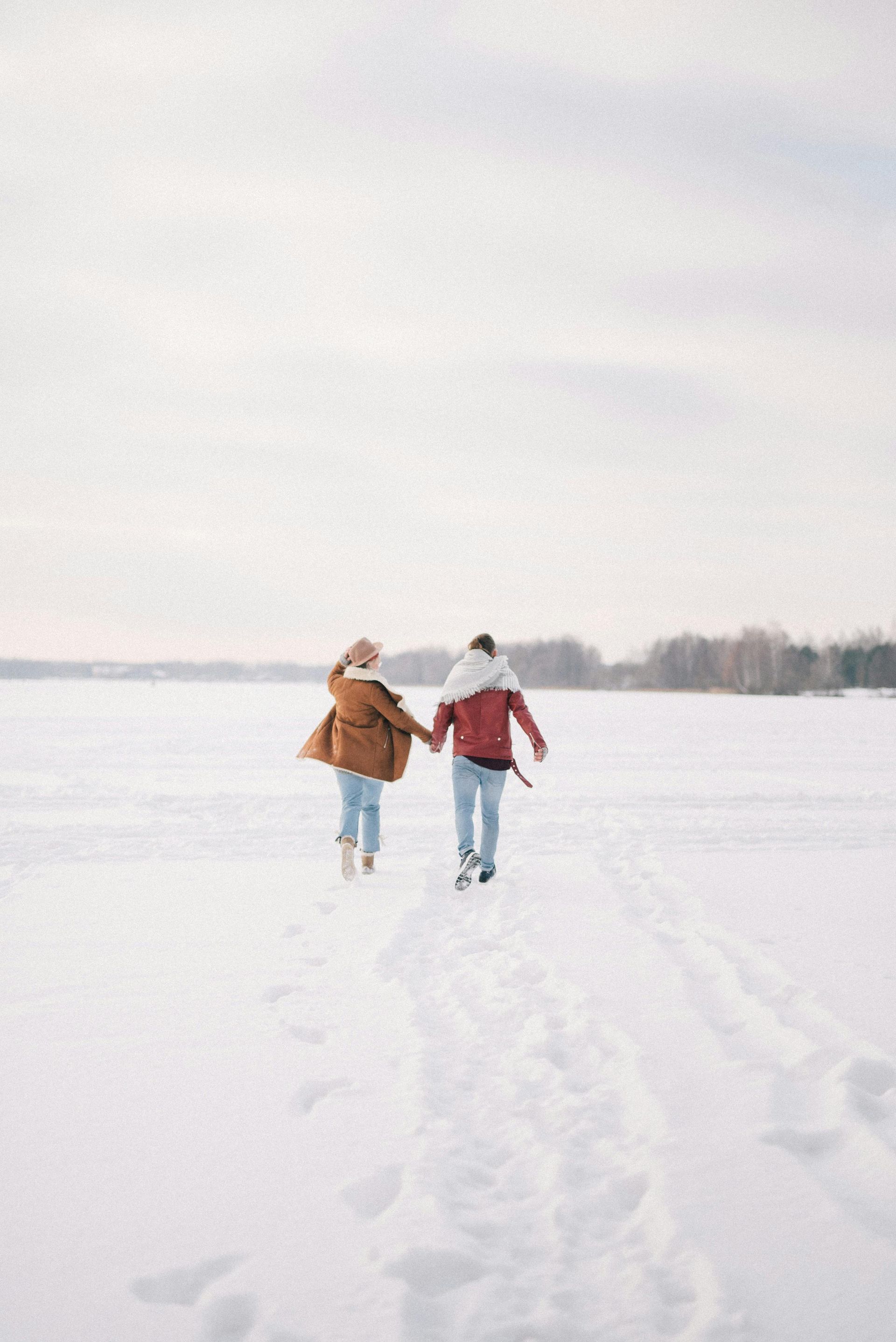 Two people holding hands, walking across a snowy landscape, leaving footprints.