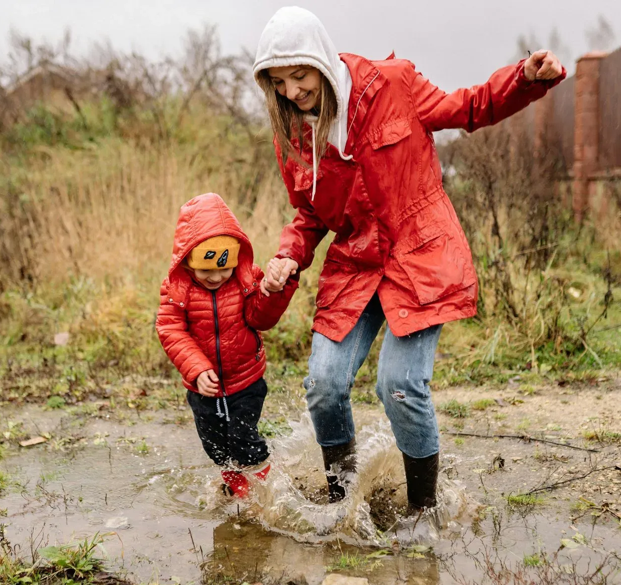 Adult and child in red raincoats splashing through a muddy puddle outdoors