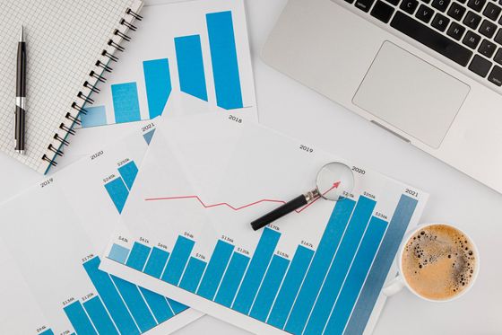 Overhead view of a work desk with charts, a laptop, notepad, pen, magnifying glass, and a cup of coffee.