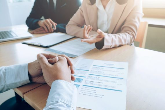 A job interview is in progress. A person's resume sits on the table while interviewers in business attire ask questions.